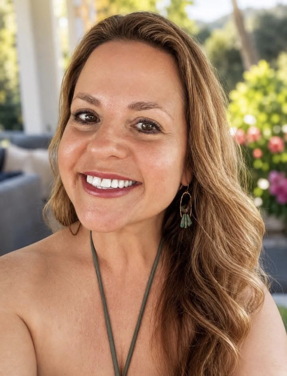Close-up of a smiling woman with long, wavy hair, wearing earrings and a necklace, standing outside with colorful flowers in the background.