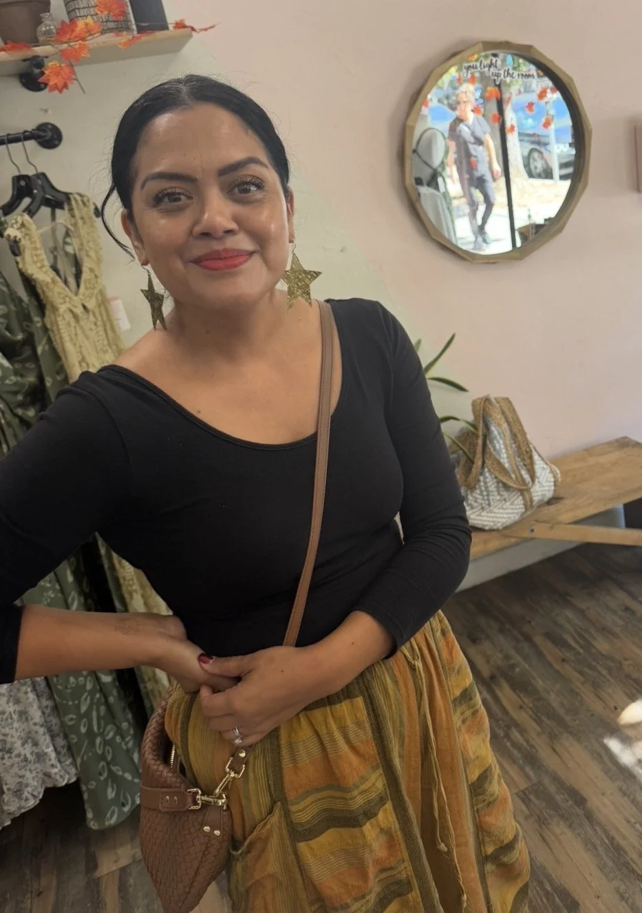 A woman with dark hair, wearing a black top and striped orange, yellow, and brown pants, standing indoors near a mirror, with a handbag hanging from her shoulder and large star-shaped earrings.