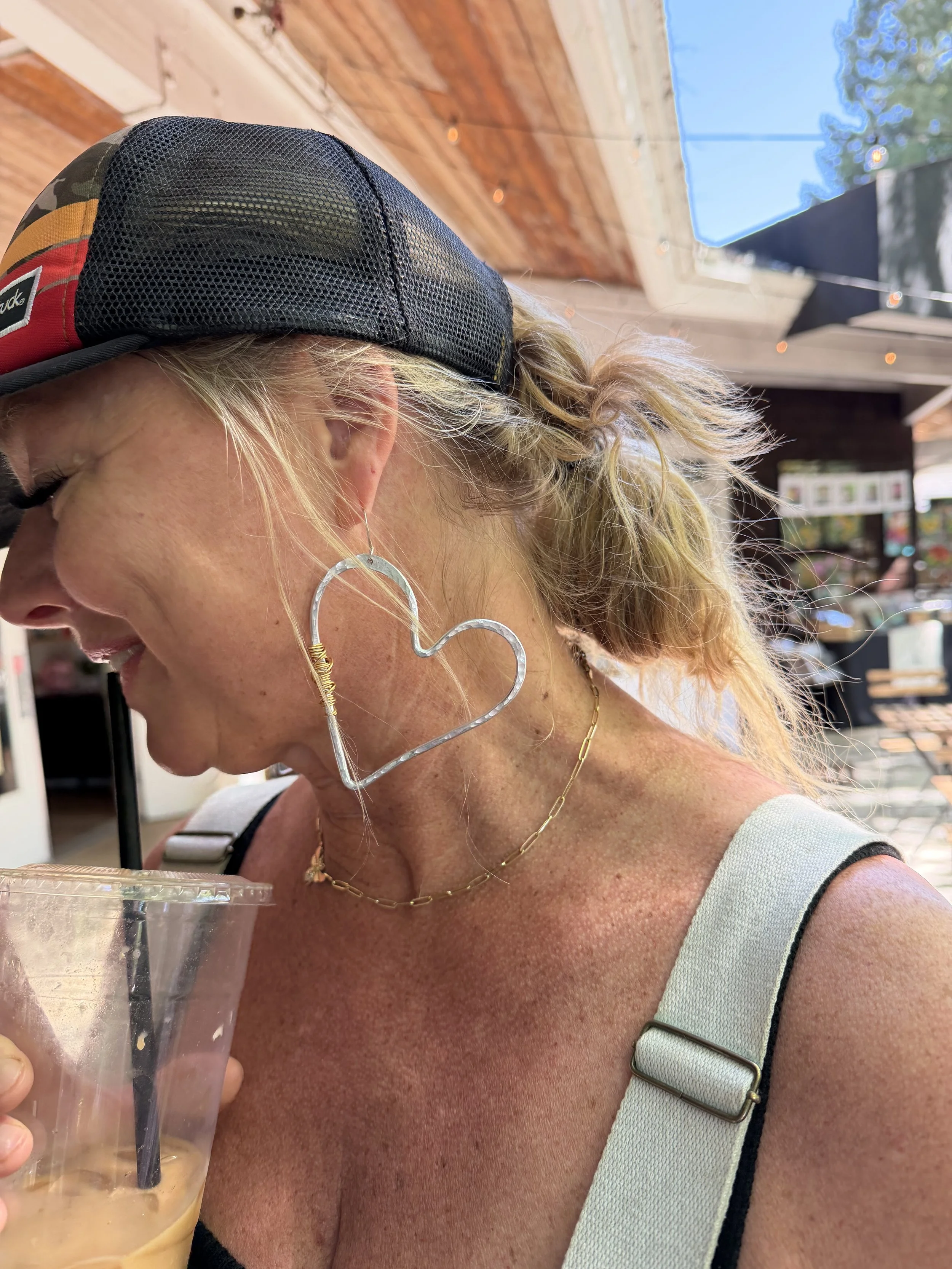 A woman with blonde curly hair wearing a black and camouflage baseball cap, hoop earrings, and a gold chain necklace, holding a clear plastic cup with iced coffee or tea. She has a tan complexion, and is wearing a light-colored tank top with black st