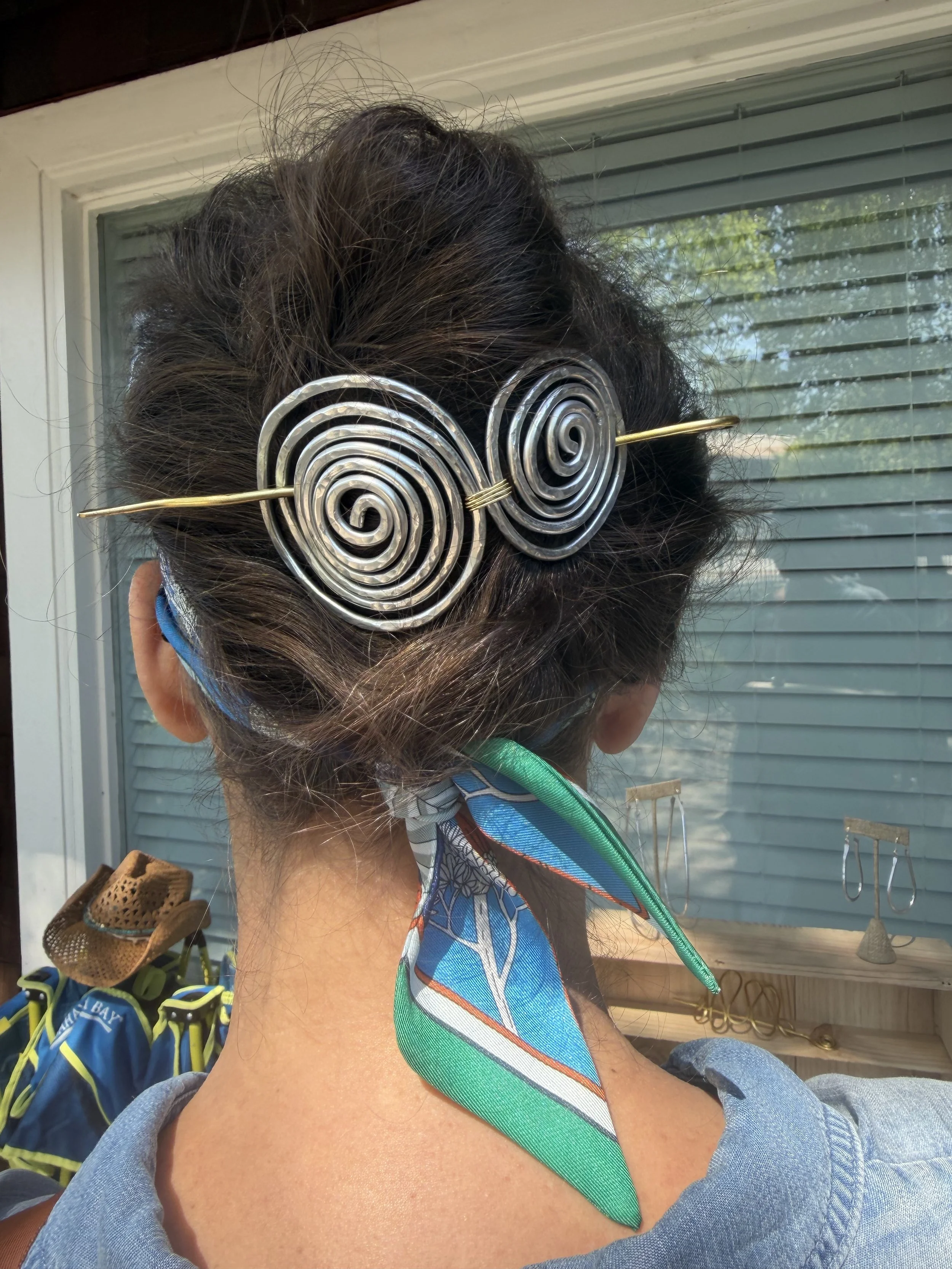 A person with short dark hair wearing a metal spiral hair accessory with a golden pin. The person is dressed in a denim jacket and is sitting outdoors in front of a window with blinds. Various items, such as colorful bags and hooks, are visible in th
