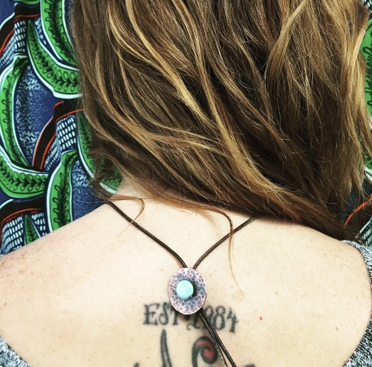 Back of a person's neck with a tattoo reading "Est. 1984" and a colorful necklace with a round pendant. The person's hair is brown with highlights and the background features a patterned fabric with green and blue designs.