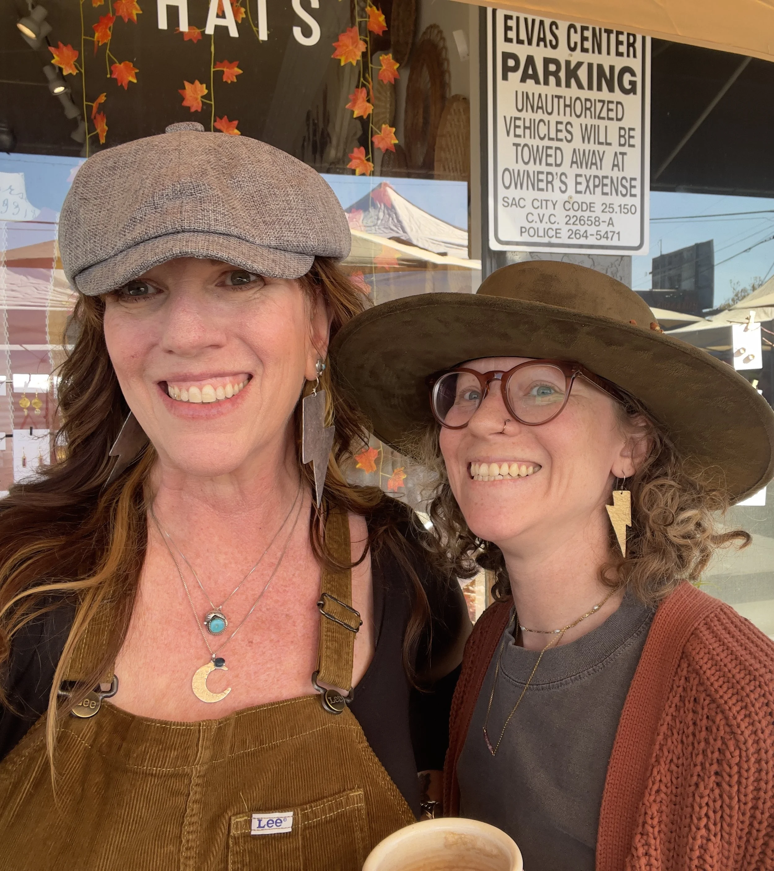 Close-up of two women smiling; one wearing a gray cap, a necklace with a turquoise pendant, and a Lee brand hat; the other wearing glasses, earrings, and a wide-brimmed hat; background shows a sign for Elvas Center parking and decorative fall leaves.