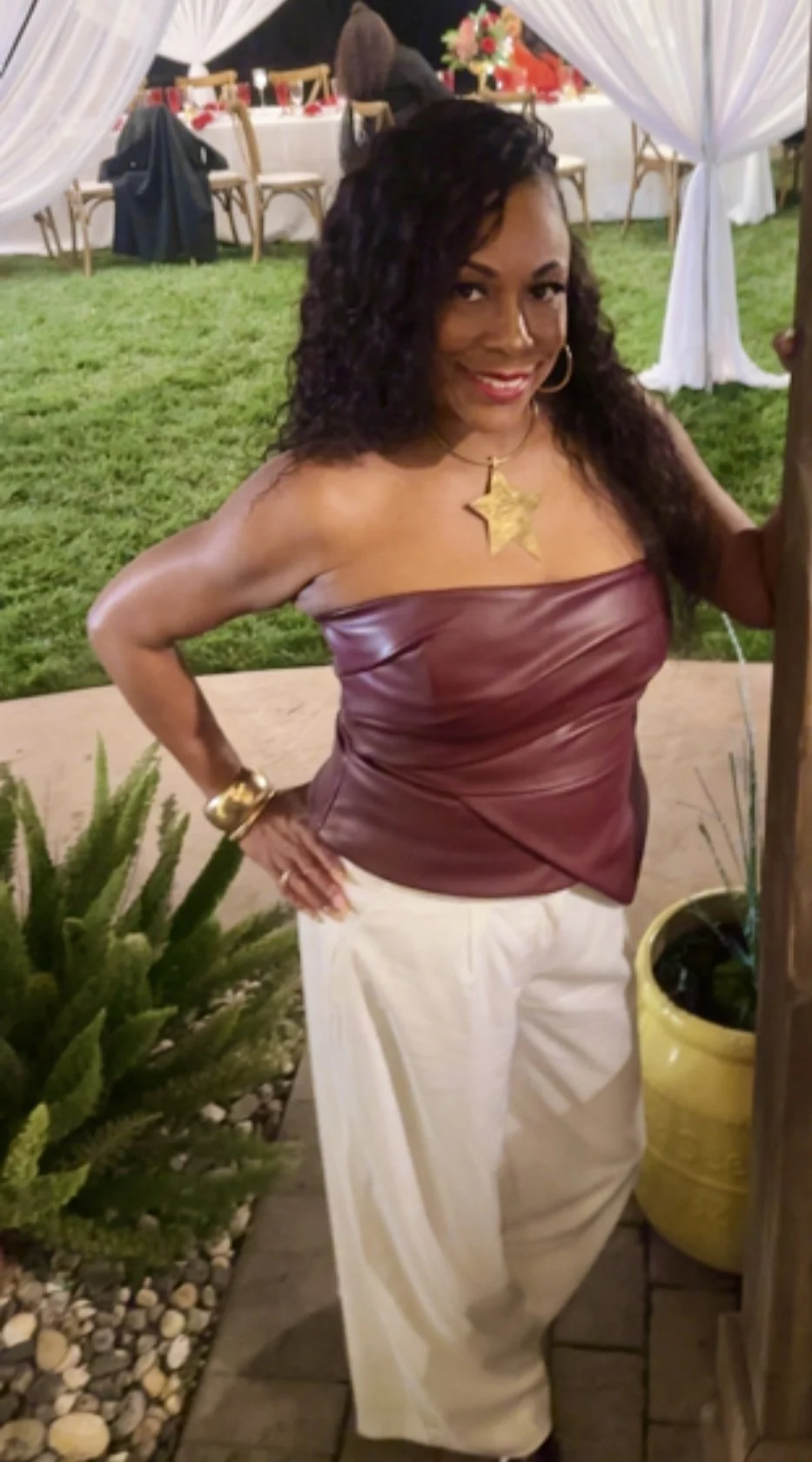 A woman standing outdoors at a decorated event, wearing a strapless maroon top, white pants, gold jewelry, and a star-shaped pendant.