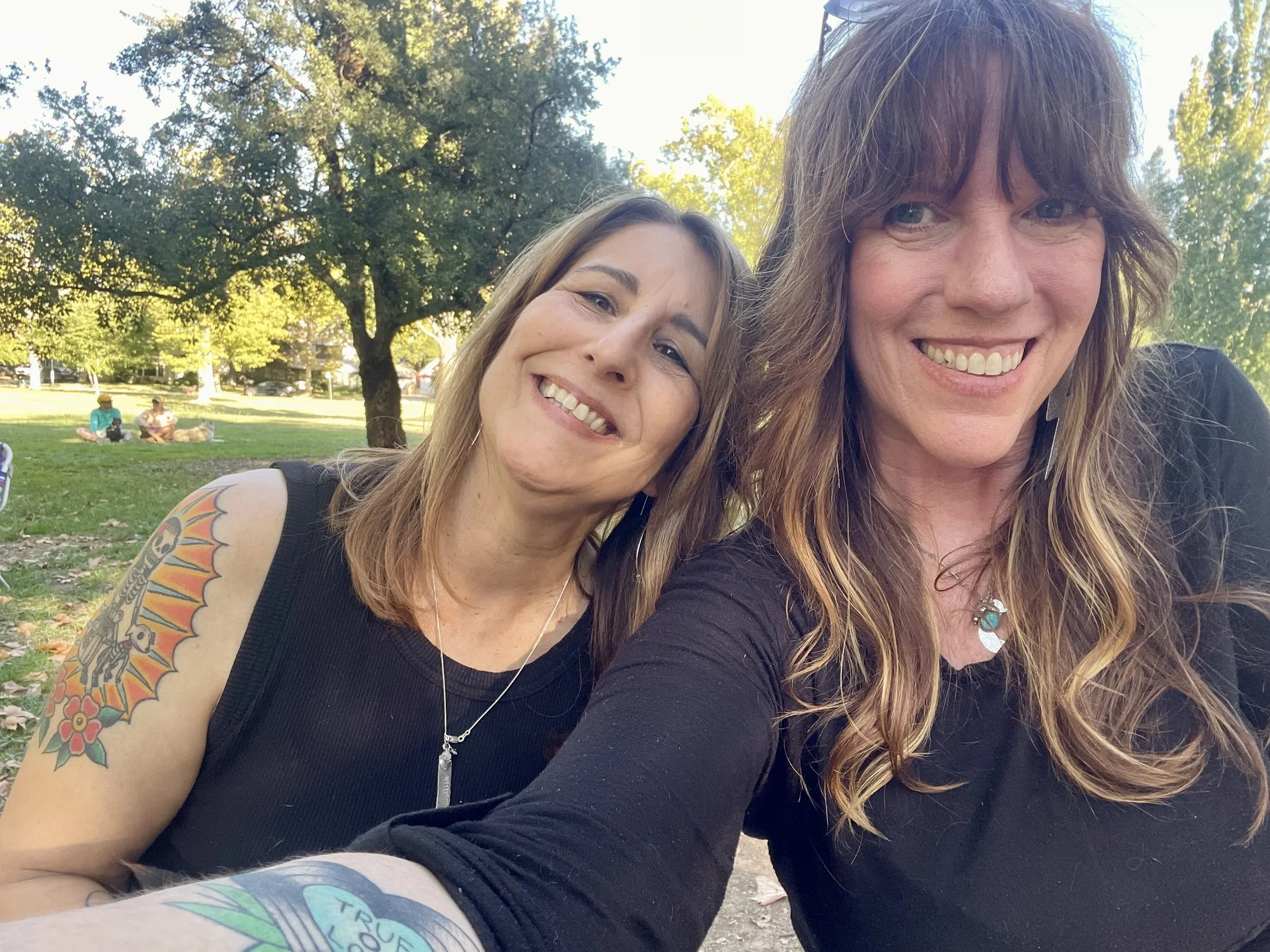 Two women taking a selfie in a park with green trees and grass in the background, smiling happily.