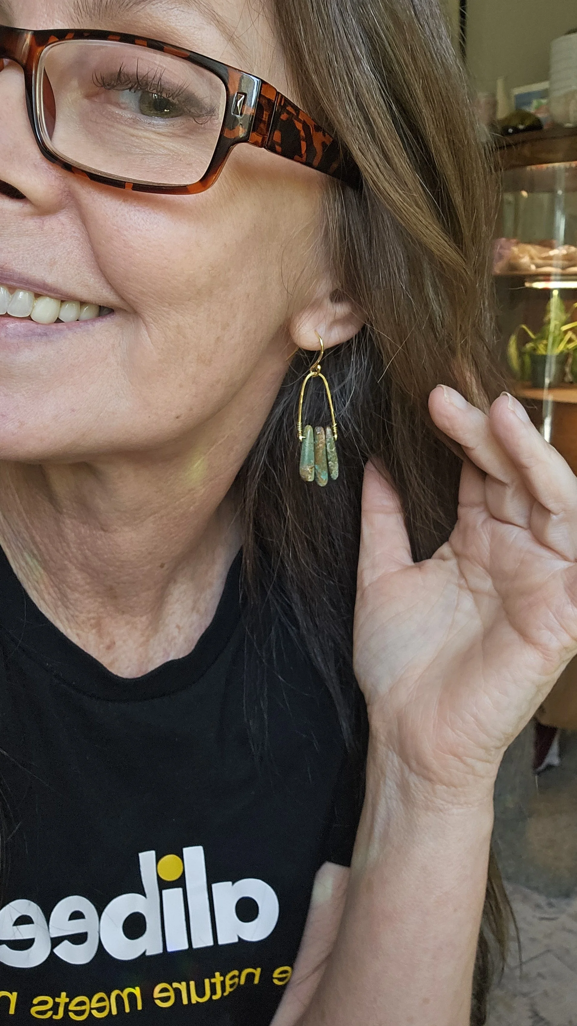 Close-up photo of a woman wearing glasses with tortoiseshell frames, golden hoop earring, smiling, and wearing a black t-shirt with yellow and grey text, holding her hair.