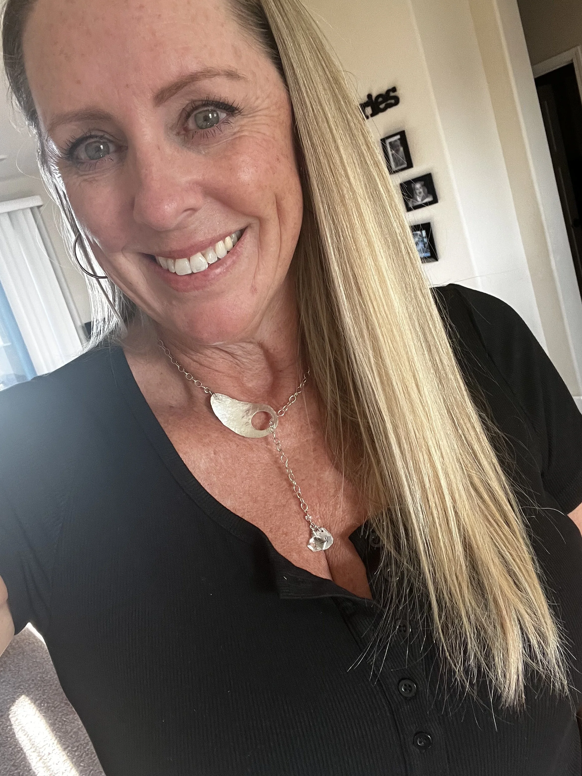 Close-up of a smiling woman with long blonde hair, wearing a black top and a silver necklace, in a well-lit room with framed photos on the wall.