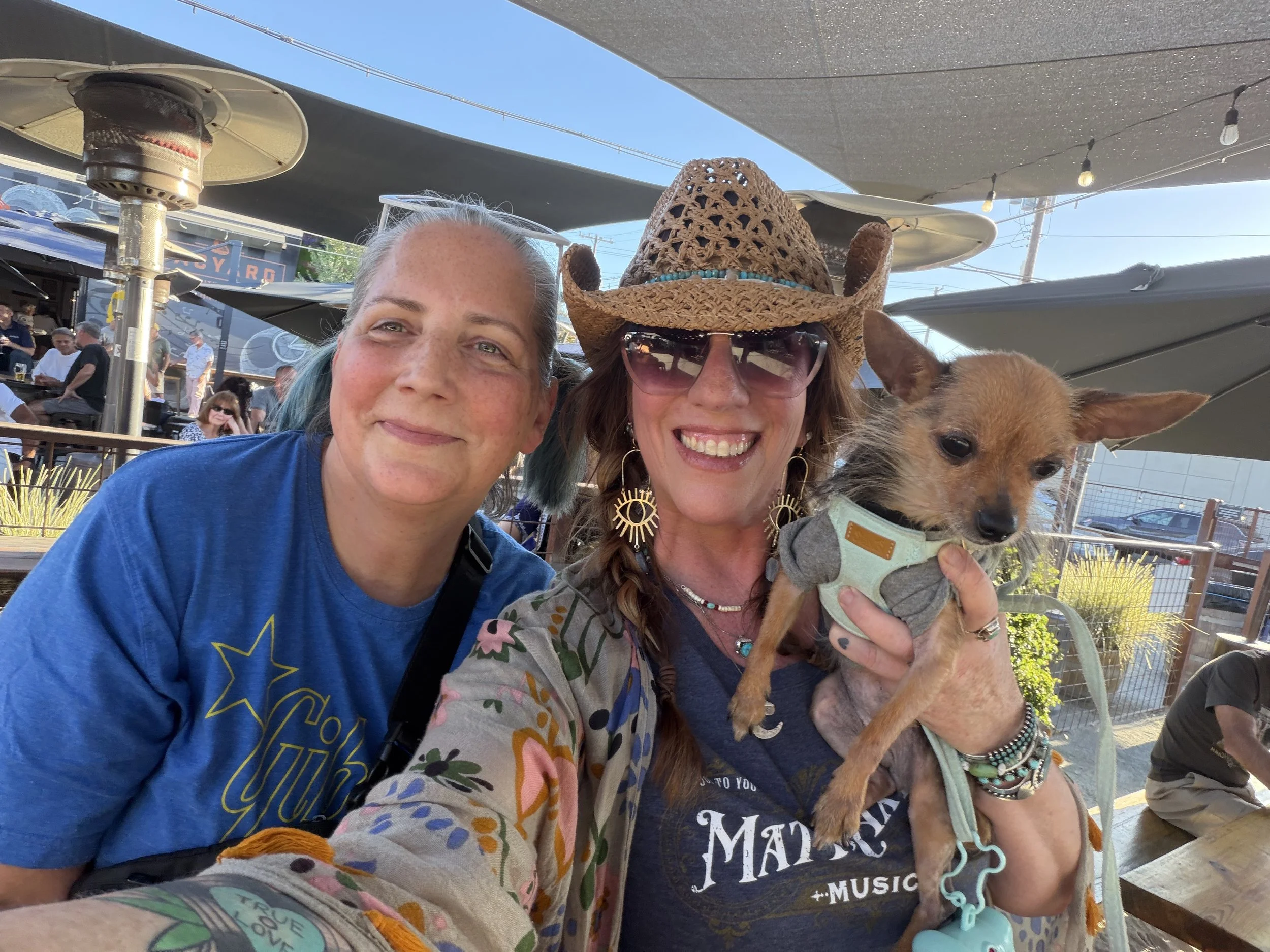 Two women smiling outdoors at a gathering, one wearing a straw hat and sunglasses, holding a small dog, with other people and outdoor setting in the background.