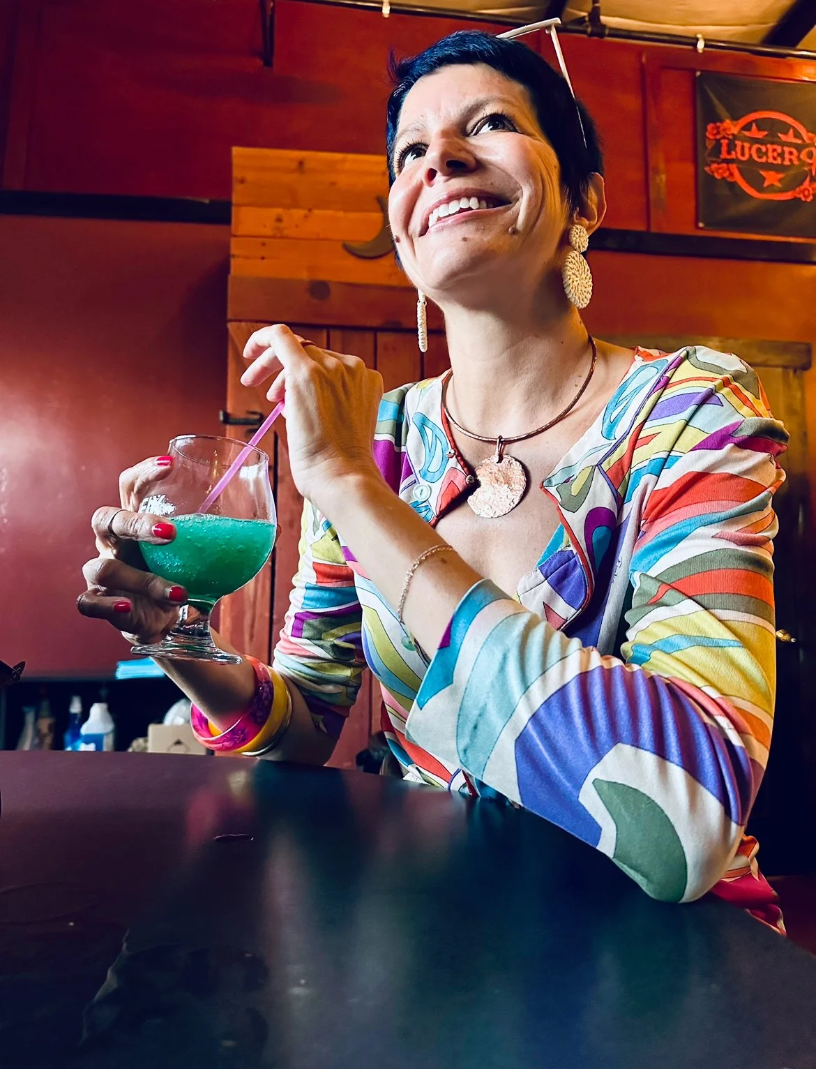 A woman with short dark hair smiling and holding a blue-green cocktail in a large round glass with a pink straw, sitting at a table in a wooden-paneled bar or restaurant.