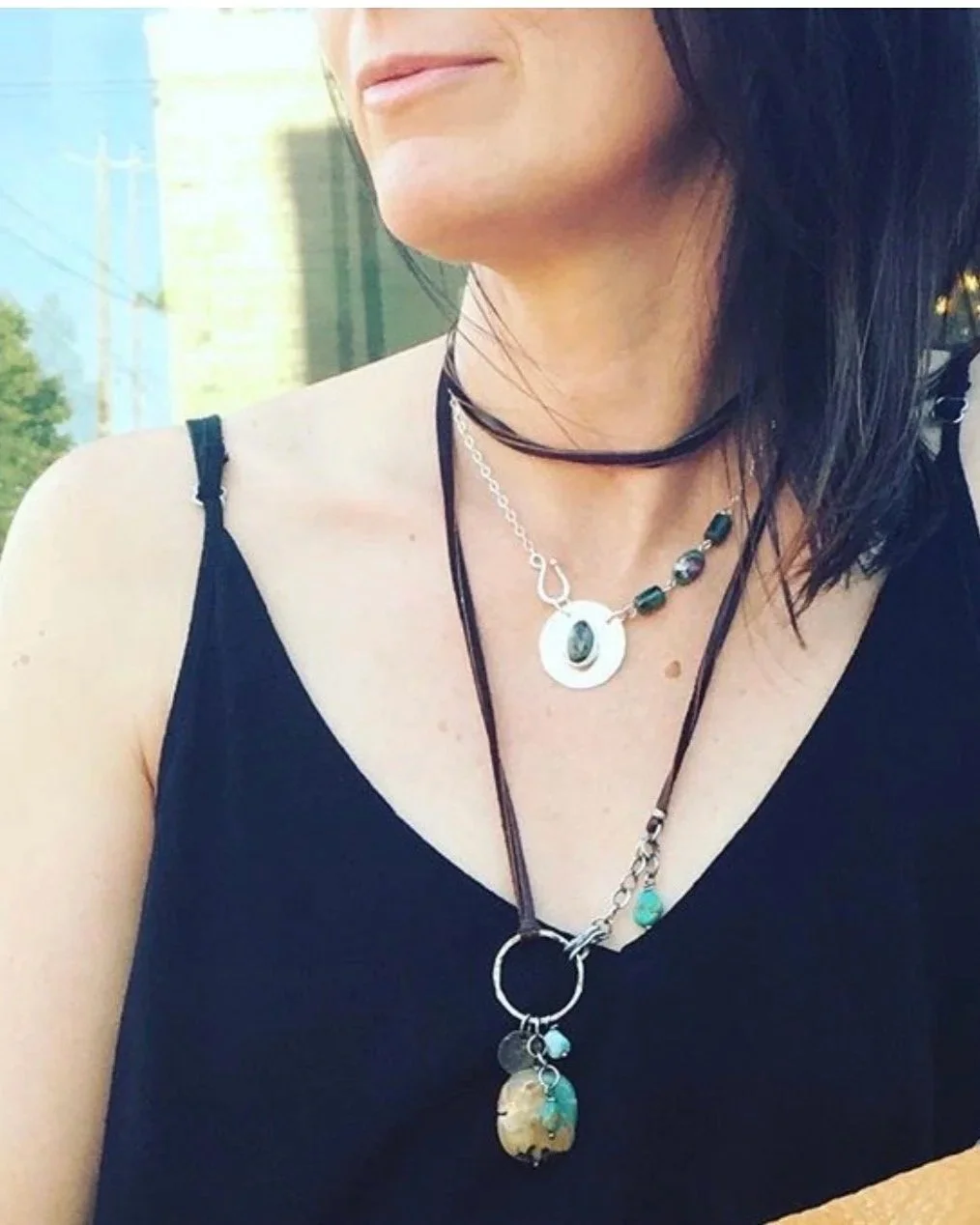 Close-up of a woman's neck and shoulders, wearing layered necklaces and a black sleeveless top, with an outdoor cityscape background.