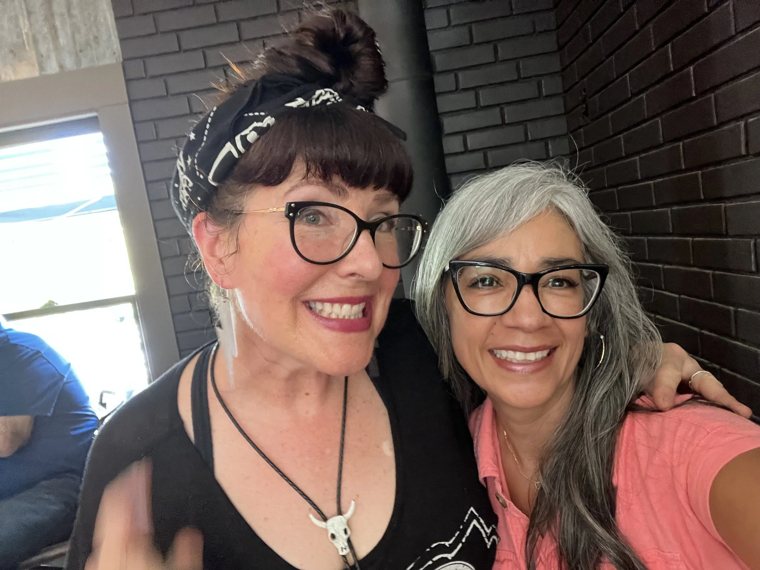 Close-up of two women smiling, sitting indoors against a black brick wall. The woman on the left has short brown hair with a hair scarf, glasses, and is wearing a black shirt. The woman on the right has gray hair, glasses, and is wearing a pink shirt