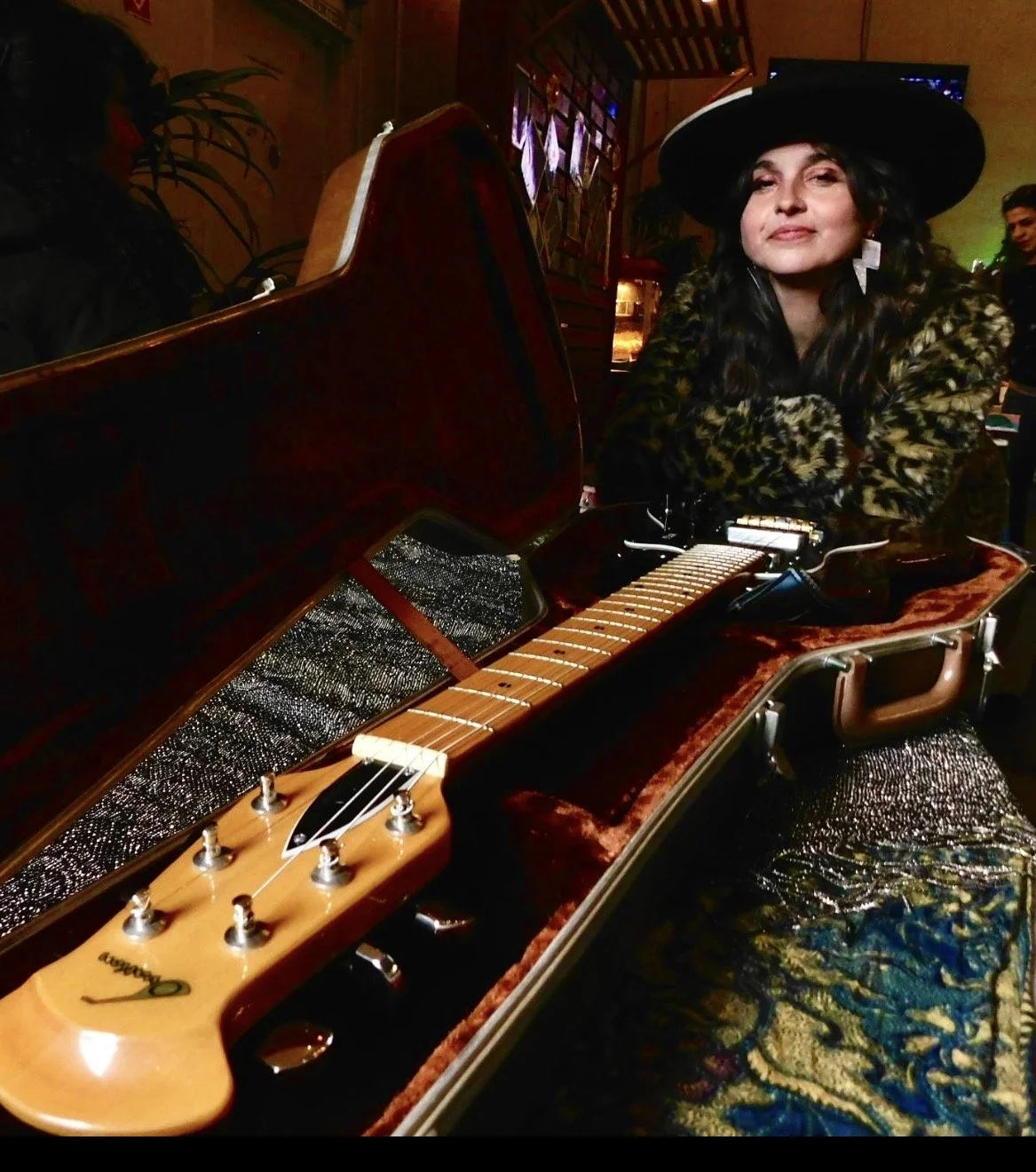 A woman with long dark hair, wearing a large black hat, a leopard print coat, and patterned skirt, sitting at a vintage open case piano with a guitar for a lid, in a warmly lit room with artwork on the wall and a man in the background.