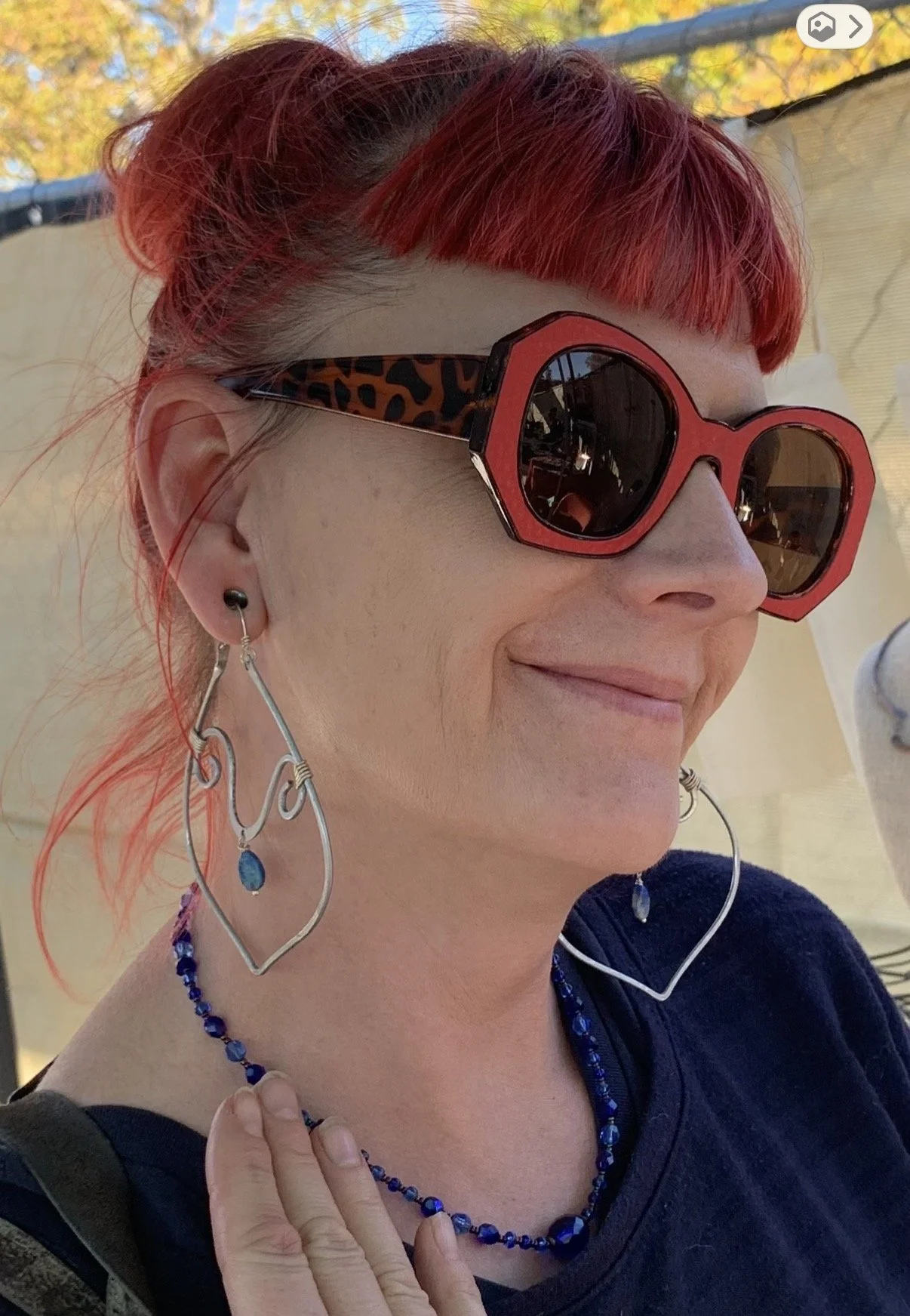 A woman with red hair and bangs wearing large red sunglasses with leopard print temples, star-shaped silver earrings with blue stones, and a blue beaded necklace, smiling outdoors in daylight.