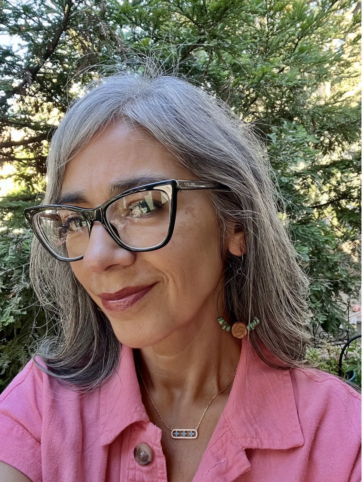 A woman with gray hair, wearing glasses, a pink shirt, and jewelry, smiling outdoors with green foliage and sunlight in the background.