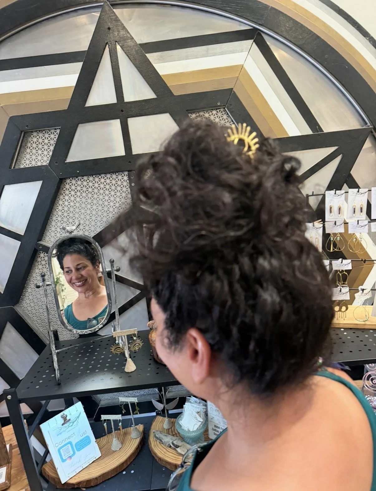 A woman with dark, curly hair and a gold hair clip looking into a small mirror at a jewelry store. The mirror reflects her smiling face. The store display includes earrings, necklaces, and other jewelry.