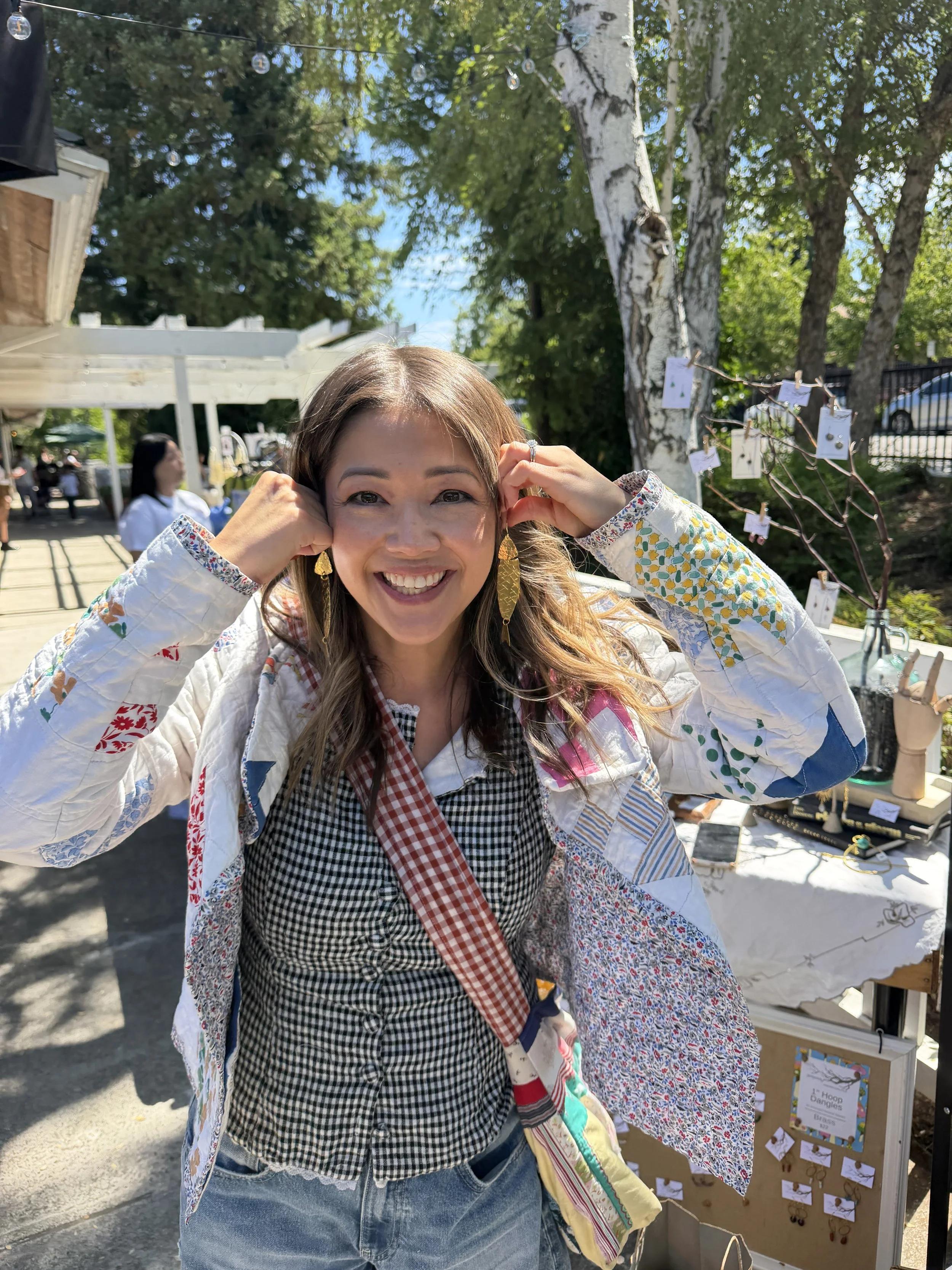 A smiling woman with long hair, wearing earrings, a checkered shirt, and a quilted jacket, stands outdoors at a market stall with jewelry displays. There are trees and other people in the background.