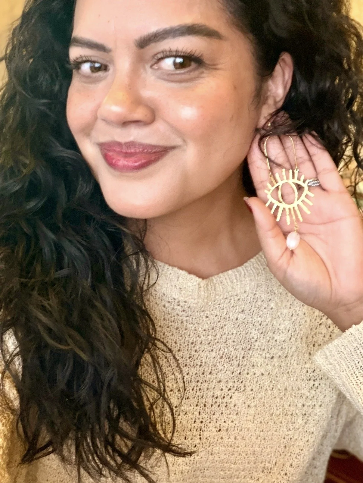 Woman with dark, curly hair holding a large gold earring with eye and sunburst design, smiling slightly, wearing a beige sweater.