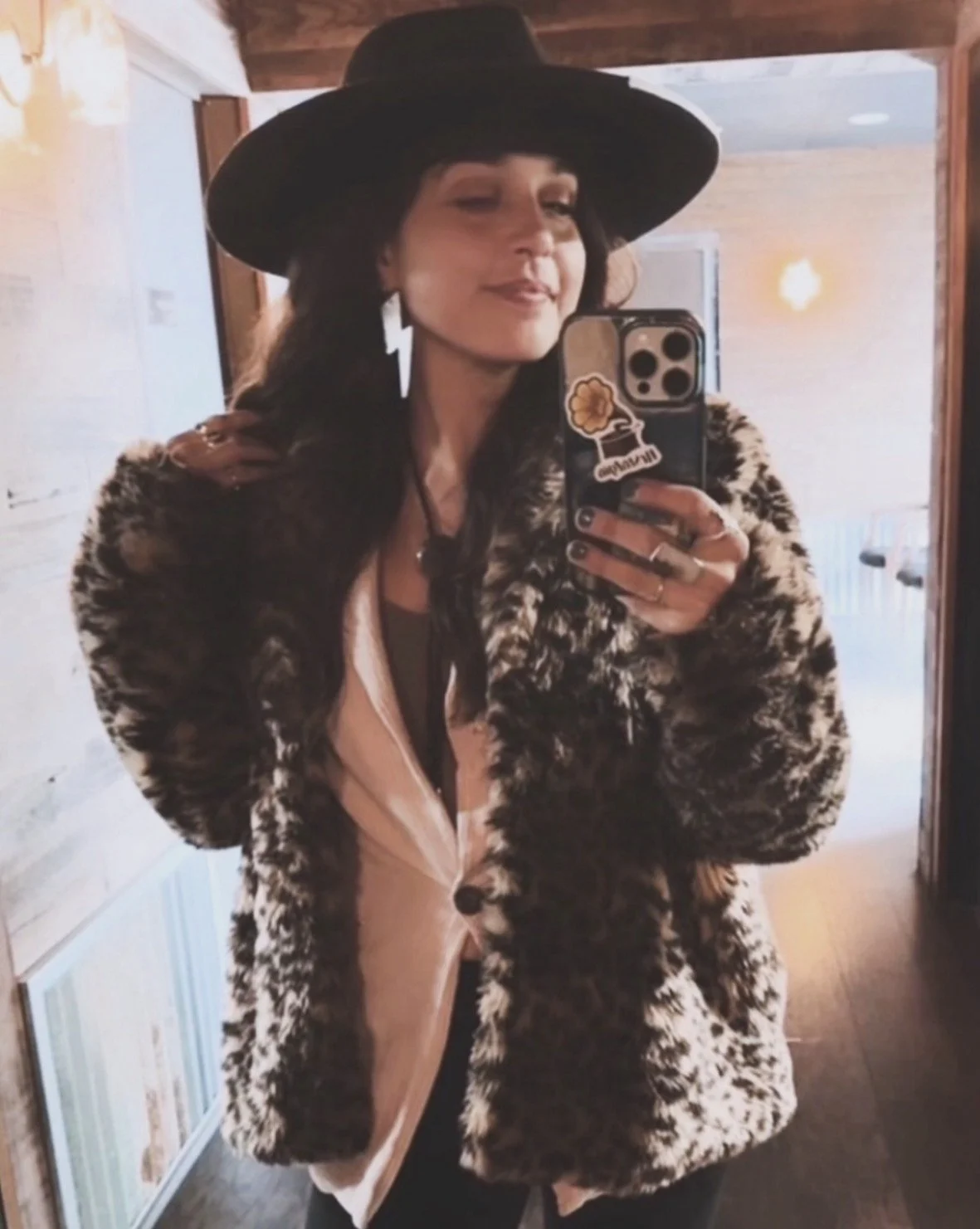 Woman taking a mirror selfie wearing a black wide-brim hat, large lightning bolt earrings, a leopard print coat, and a peach-colored hoodie.