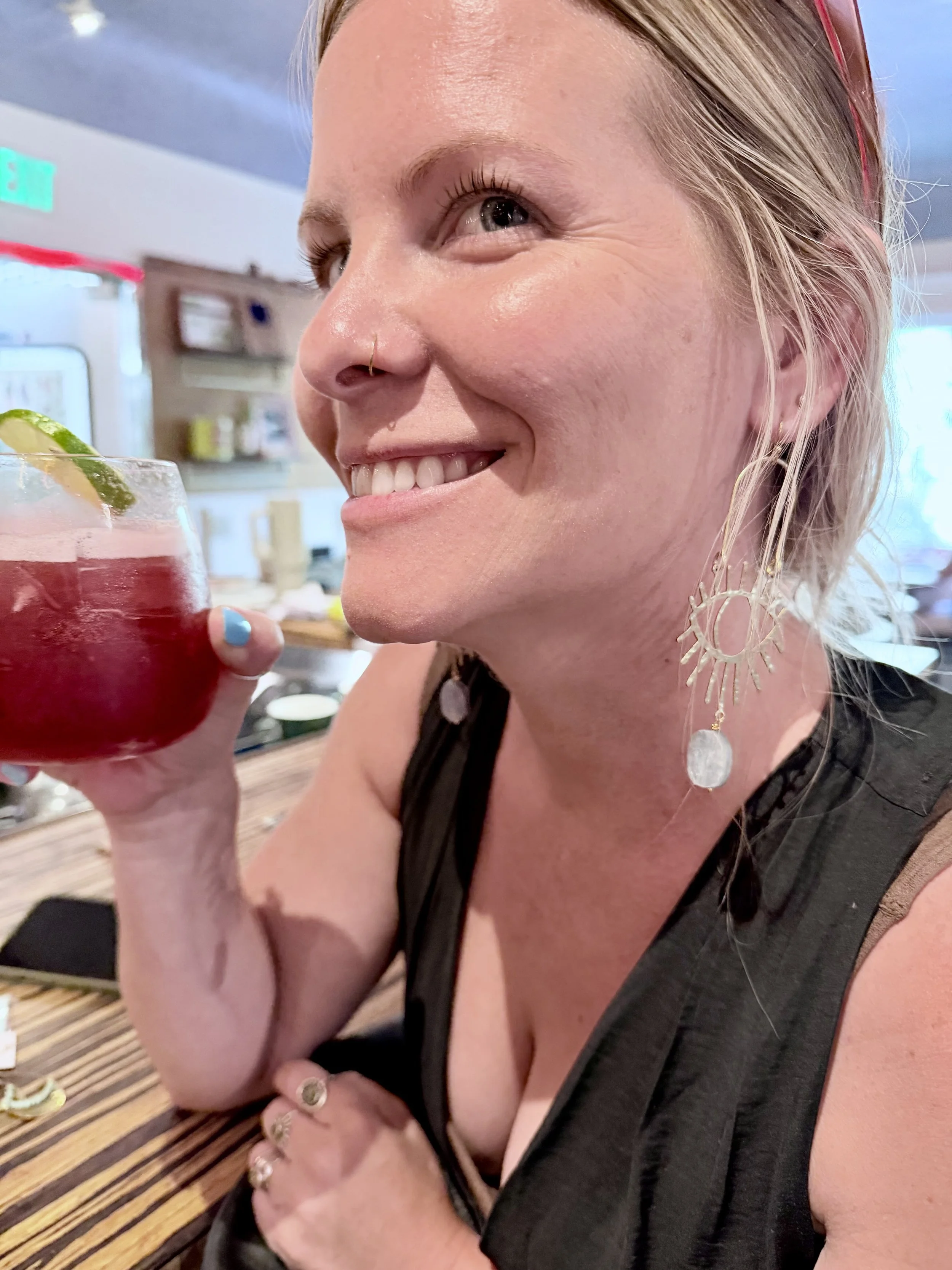 A woman with blonde hair and large decorative earrings smiling and holding a drink with a lime wedge in a restaurant or bar setting.