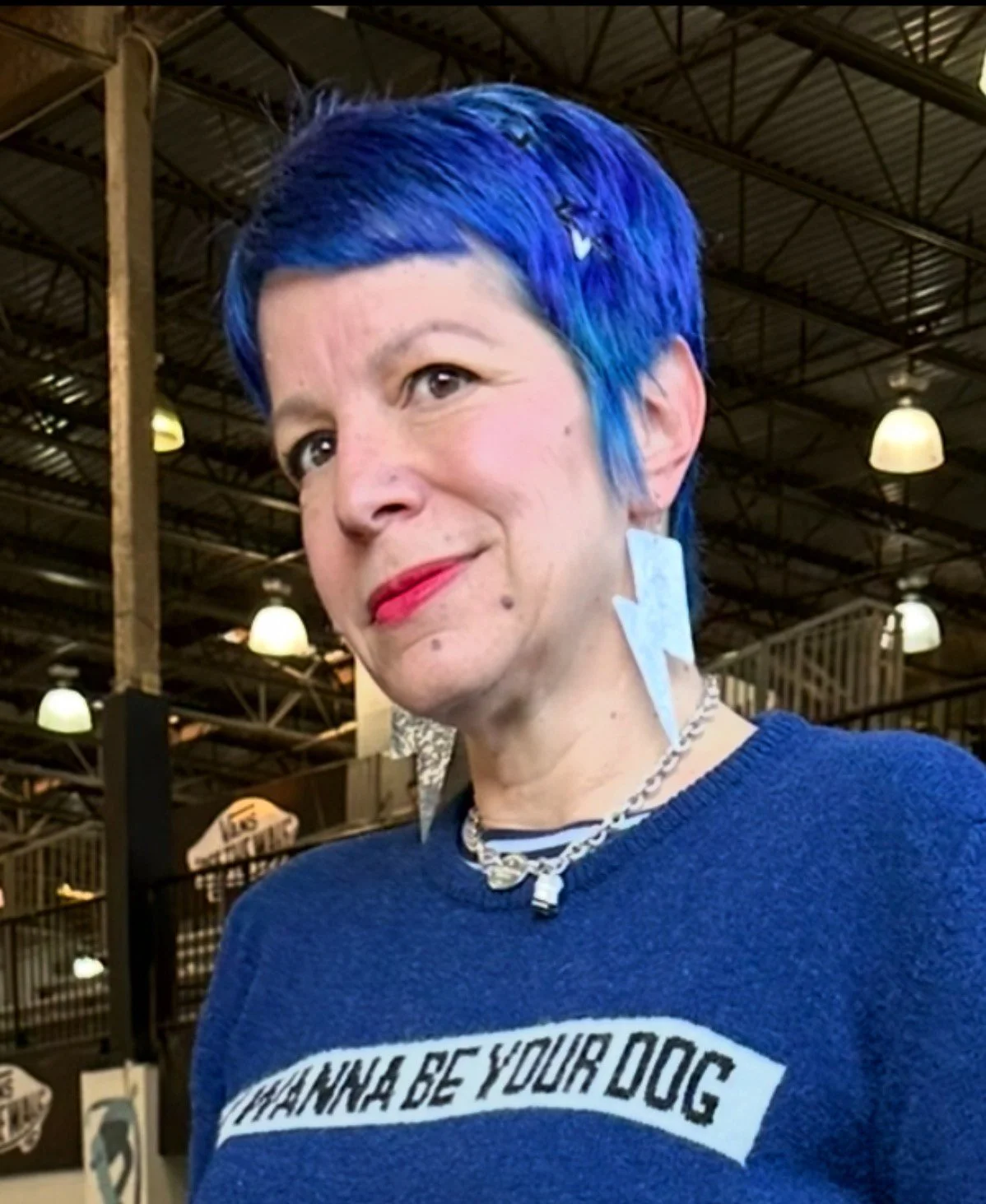 Woman with short blue hair, wearing a blue sweater with text, silver necklace, and white earrings, indoors with high ceiling and hanging lights.