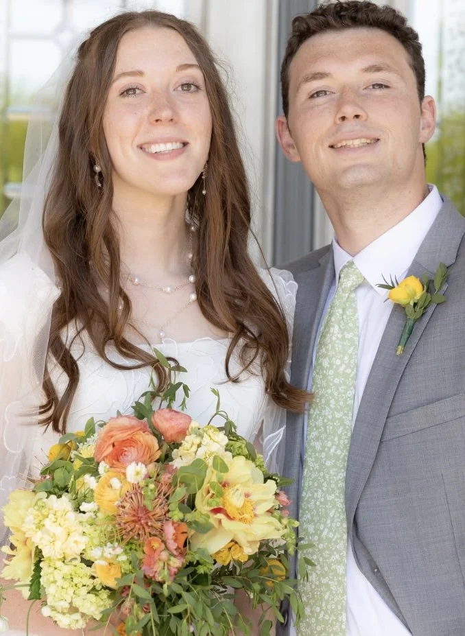 Smiling bride and groom at wedding, woman in wedding dress holding a colorful bouquet, man in gray suit with light green tie and yellow boutonniere, standing outdoors.