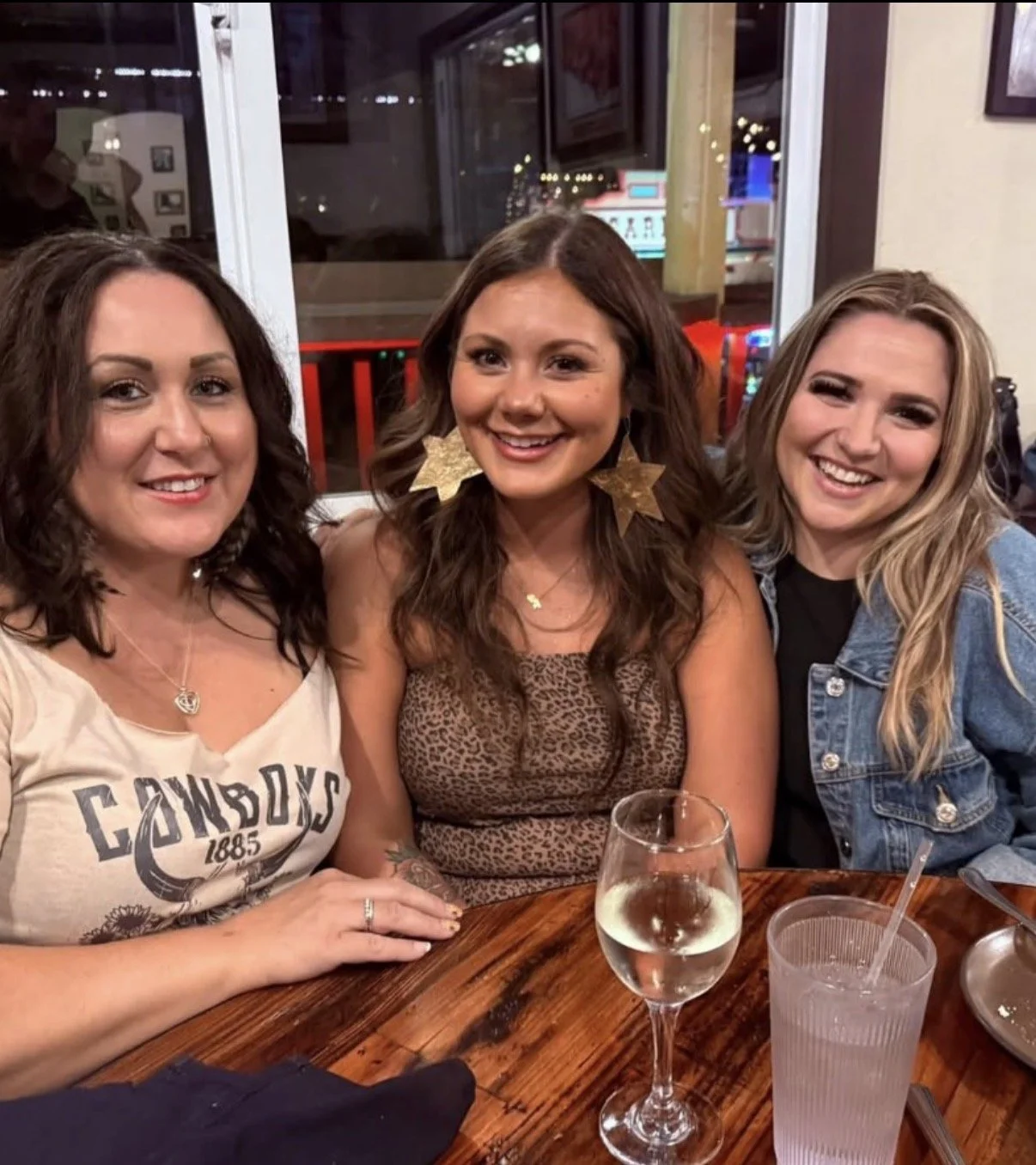 Three women sitting together at a restaurant table, smiling for the camera. The woman on the left has dark curly hair, wearing a beige shirt with text and jewelry. The woman in the center has long brown hair, wearing a leopard print top, large star-s