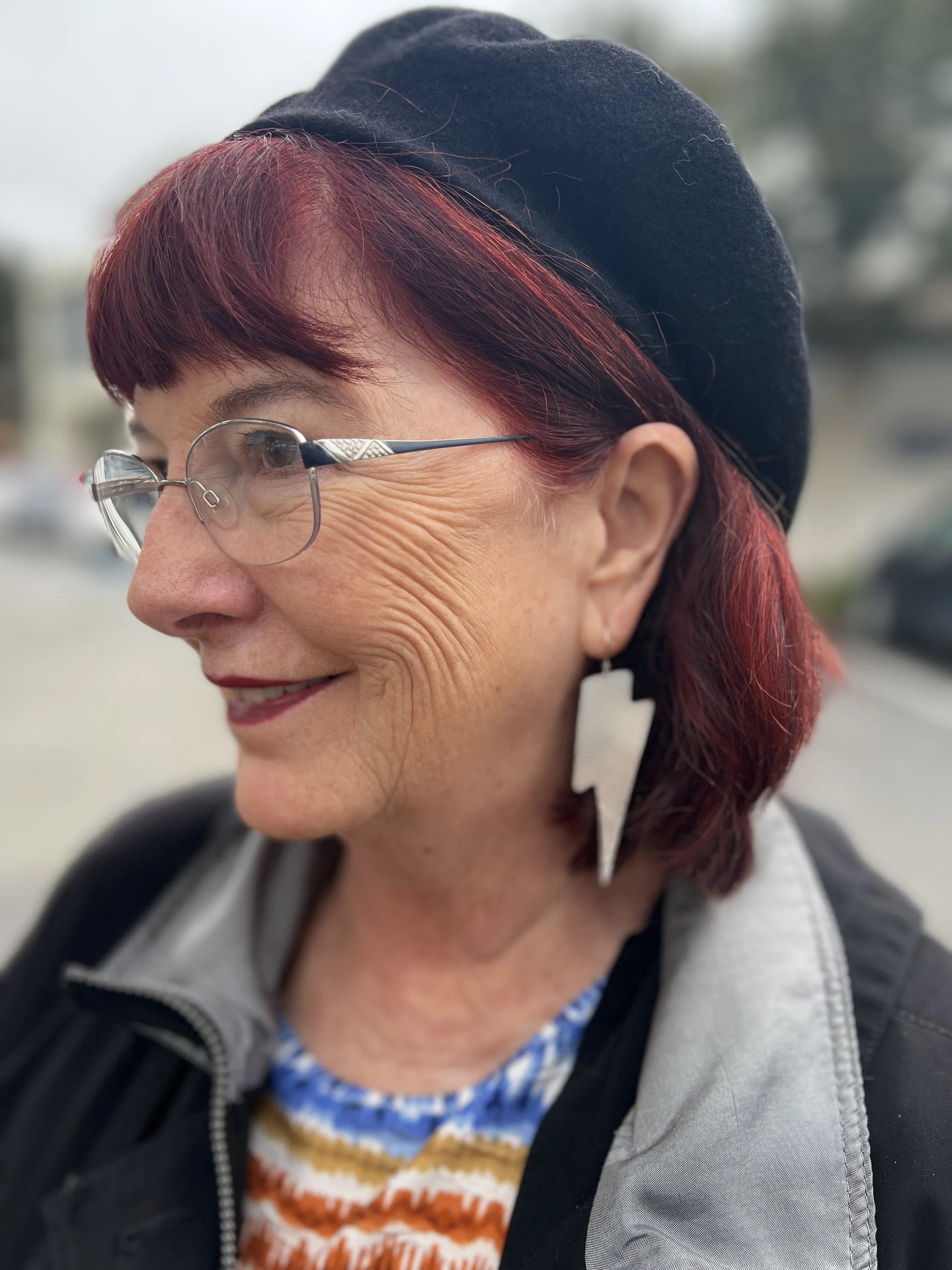 Close-up of a woman with red hair, wearing glasses, a black beret, and a large lightning bolt-shaped earring, smiling outdoors.
