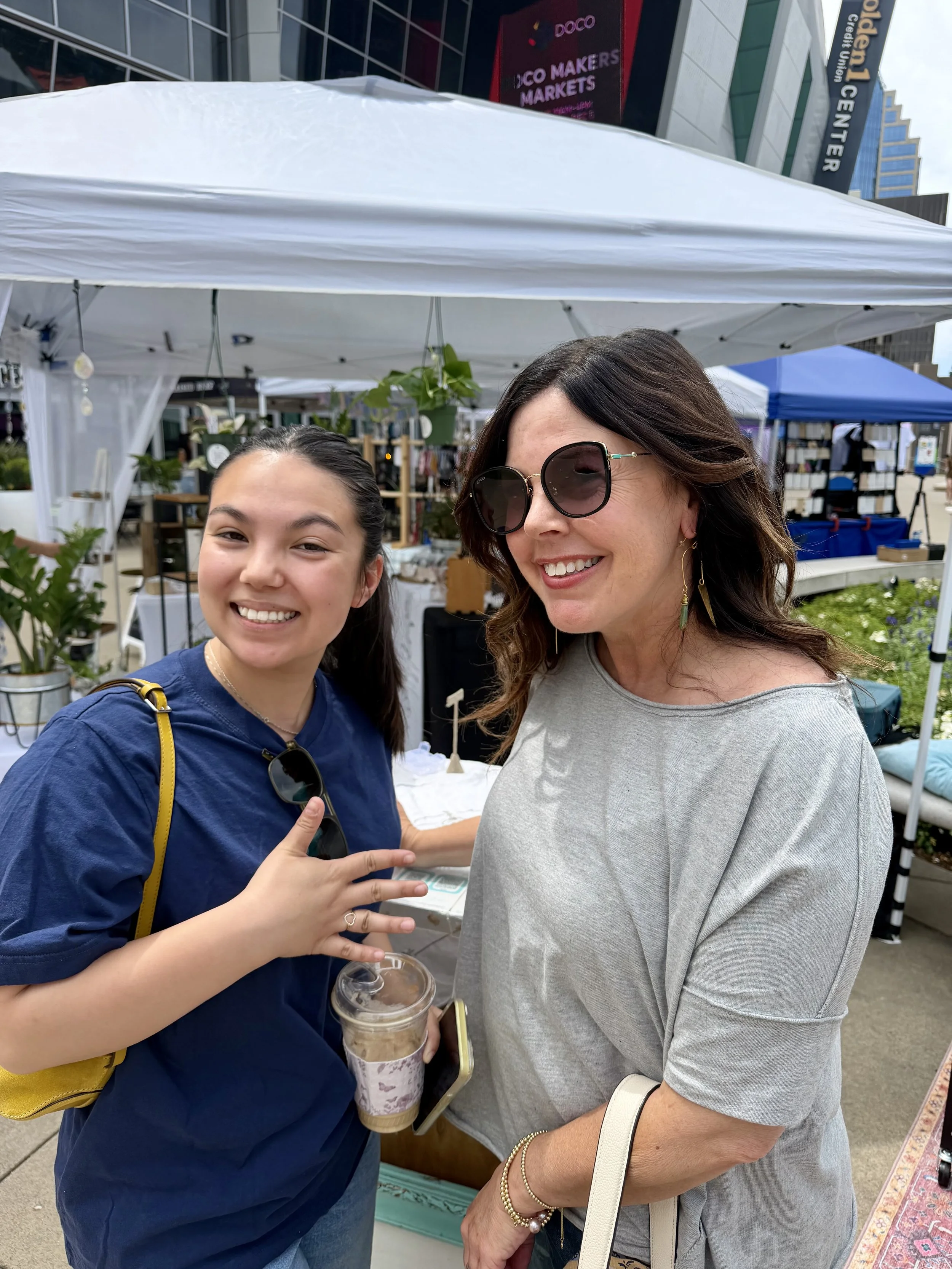 Two women smiling at an outdoor market in front of a white tent, one woman wearing sunglasses and holding a drink, the other woman pointing at her.
