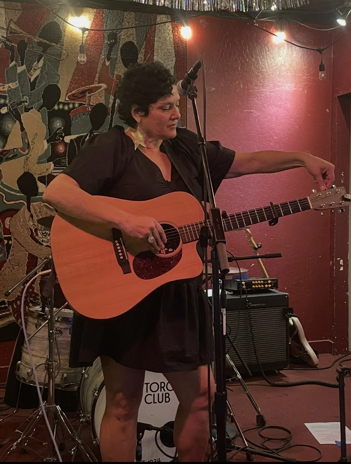 A woman with short dark hair playing an acoustic guitar on stage at the Torch Club, with colorful wall art and hanging lights in the background.
