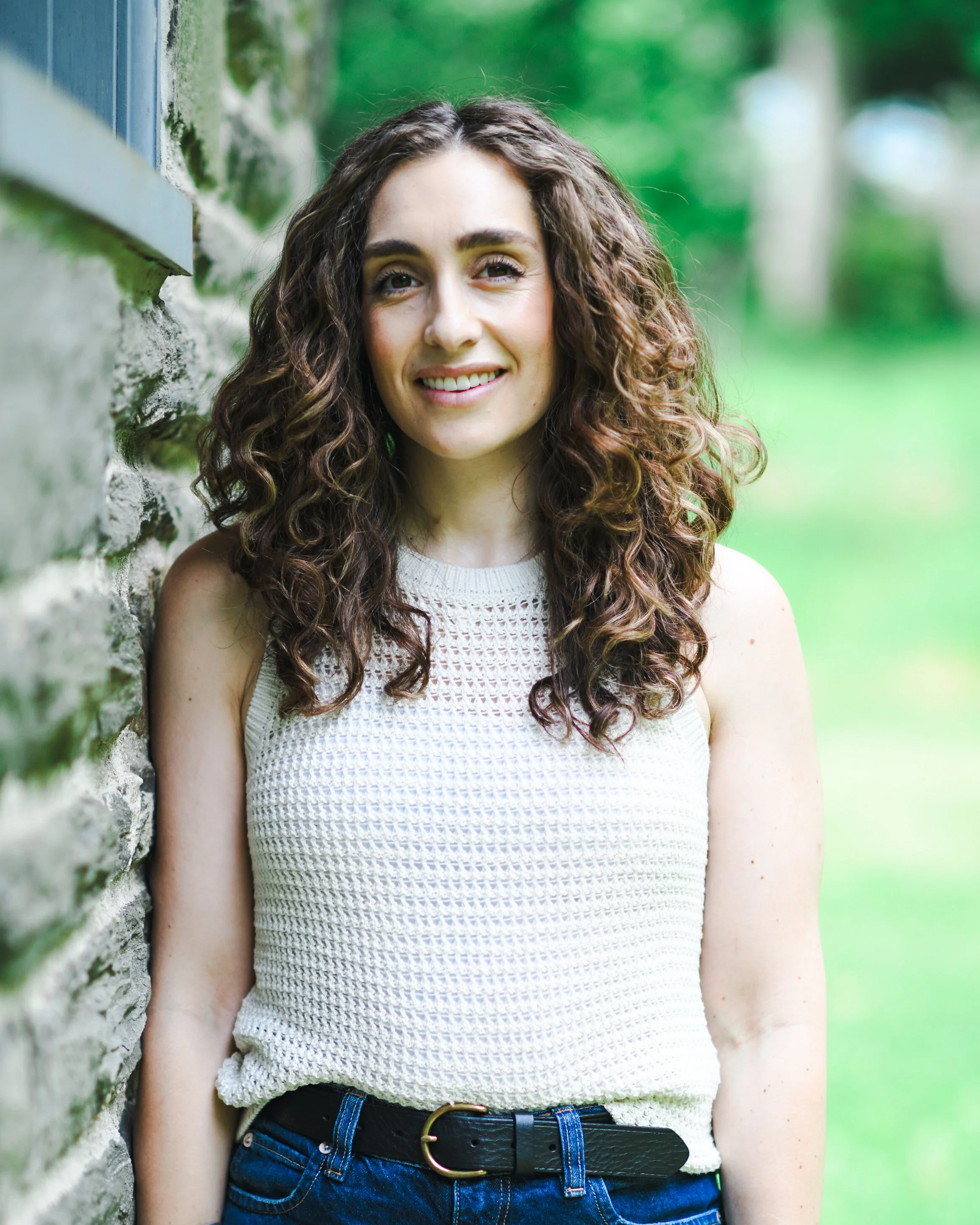 Close-up portrait of a young woman with curly brown hair, smiling, standing outside near a brick wall with greenery in the background.