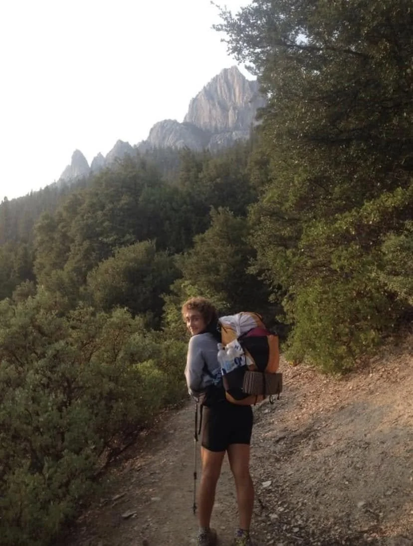 A woman hiking on a dirt trail surrounded by green trees with mountain peaks in the background, smiling and looking back.