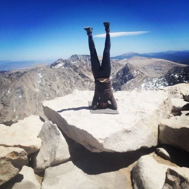 Person doing a handstand on a large rock at the summit of a mountain with rugged terrain and distant mountain ranges under a clear blue sky.