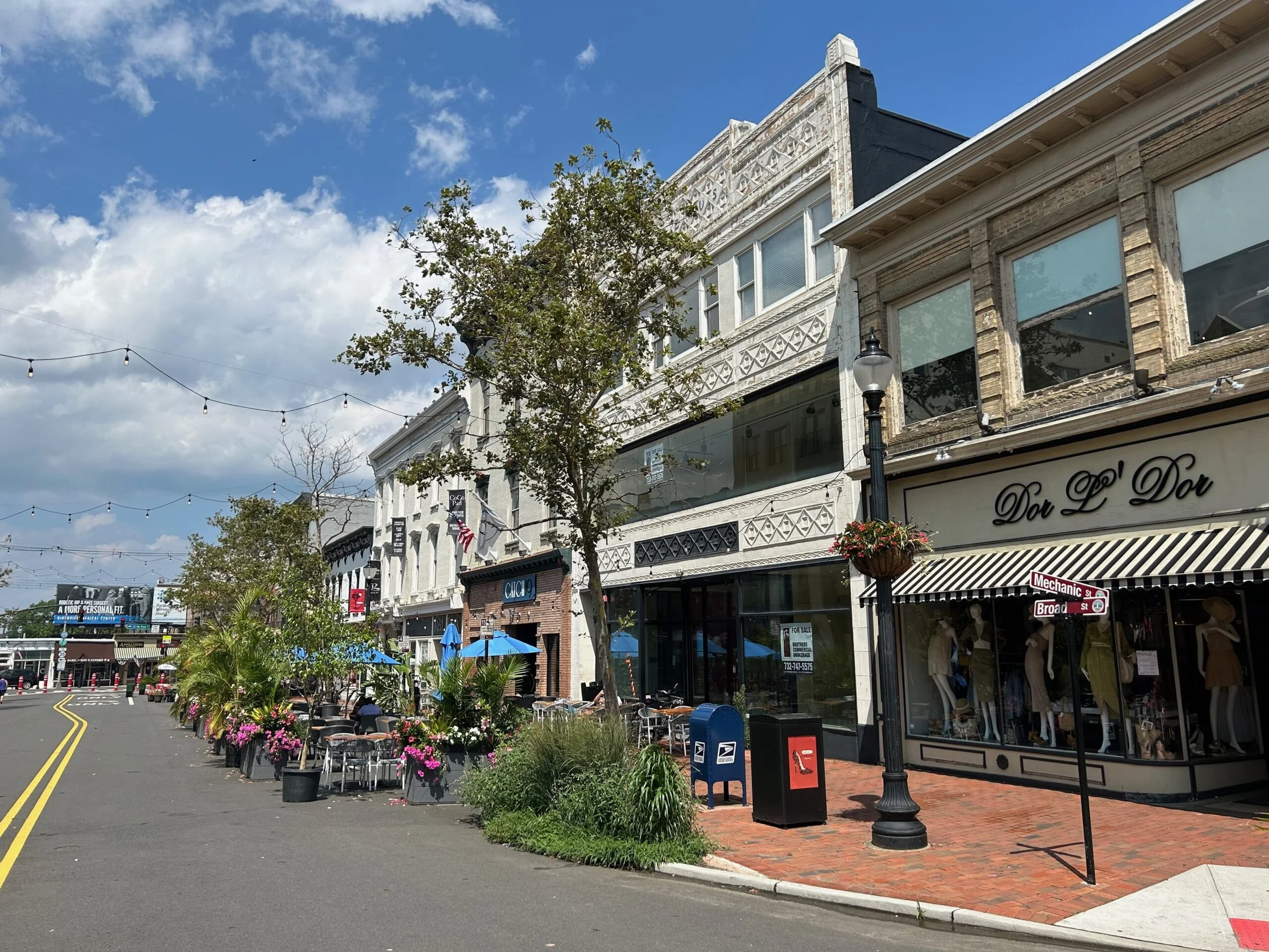 A street view of outdoor cafe seating with blue umbrellas, potted plants, and storefronts including a clothing store named Dou L D'Or, under string lights on a sunny day with a partly cloudy sky.