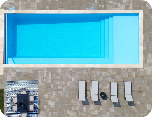 Overhead view of a swimming pool with four lounge chairs, a small round table, and a striped towel with shoes and bag on it next to the pool.