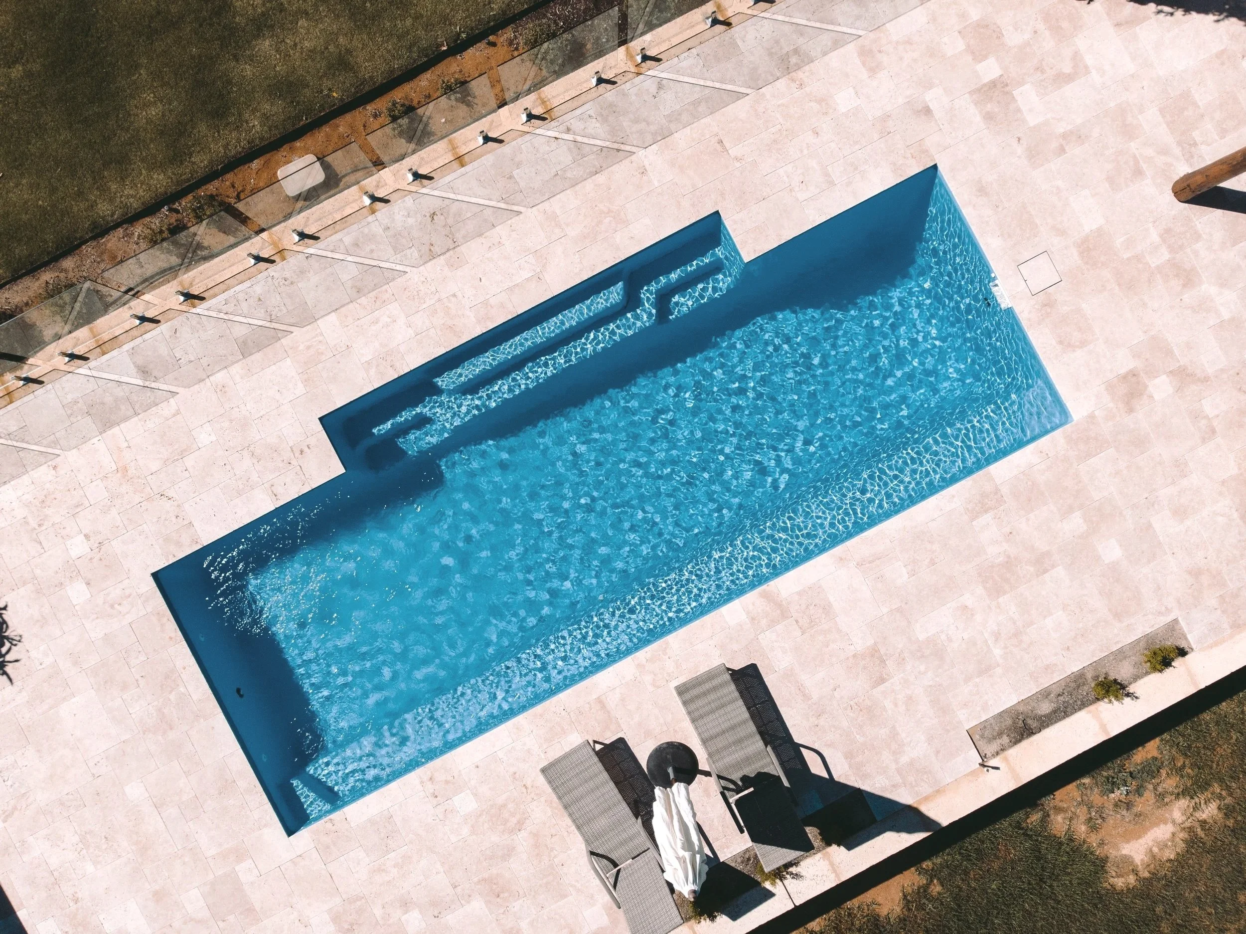 A clear blue rectangular swimming pool with steps leading into the water, surrounded by light-colored tiled pavement, with black lounge chairs and a few small plants nearby.