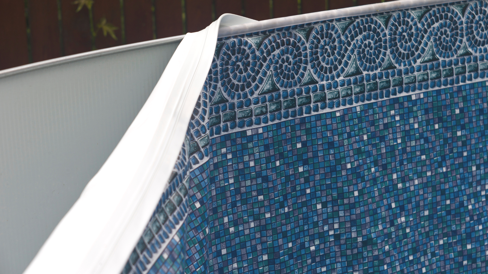 Close-up of a swimming pool wall with blue mosaic tiles and part of a white privacy fence in the background.