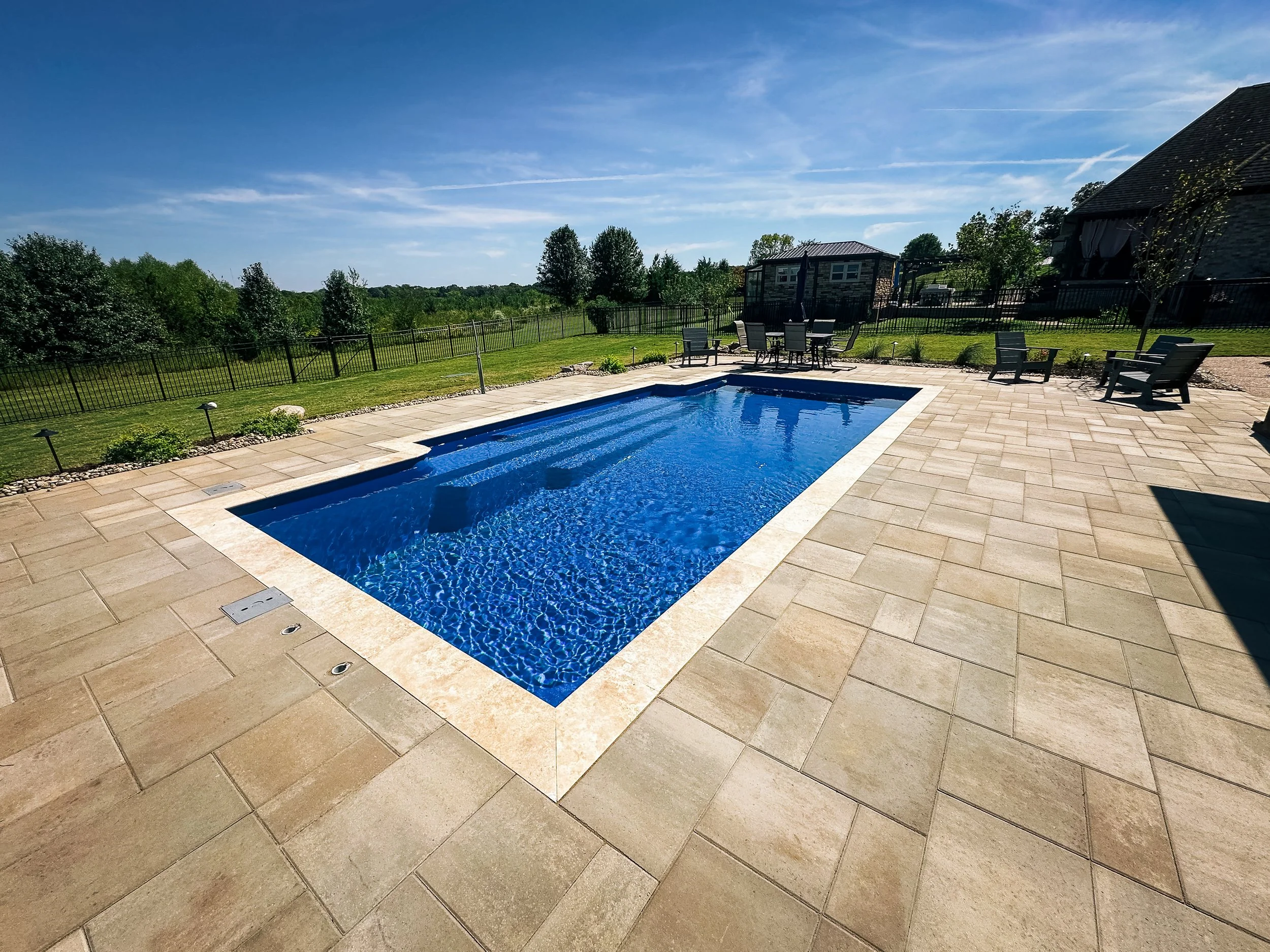 A backyard with a rectangular swimming pool, surrounding stone patio, outdoor table and chairs, fenced yard, and house with a sloped roof in the background under a blue sky.