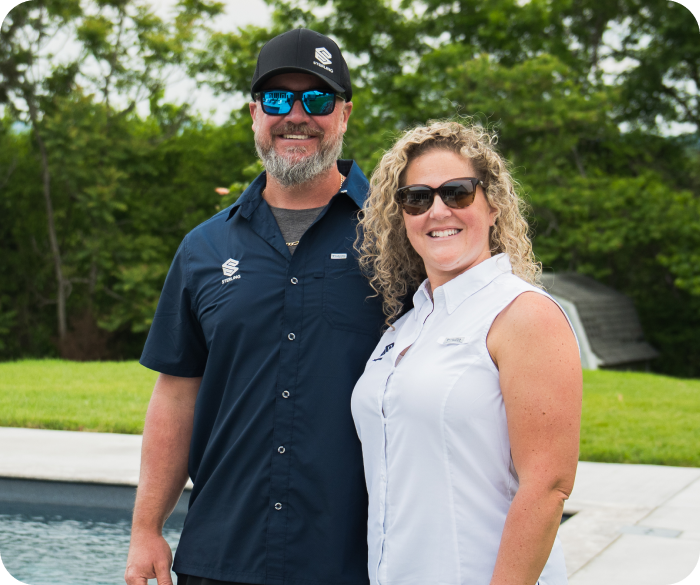 A man and woman stand outdoors by a pool, smiling and wearing sunglasses, with trees in the background.