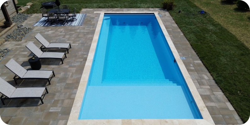 Above ground rectangular swimming pool with clear blue water, surrounded by a paved patio area with lounge chairs and a dining table with chairs.