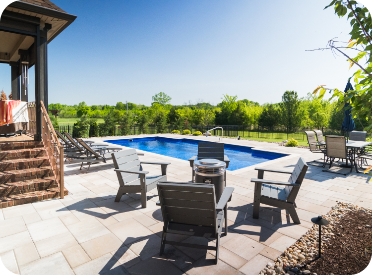A backyard scene with a swimming pool, tan patio chairs, a table with an umbrella, and a green landscape in the background under a blue sky.