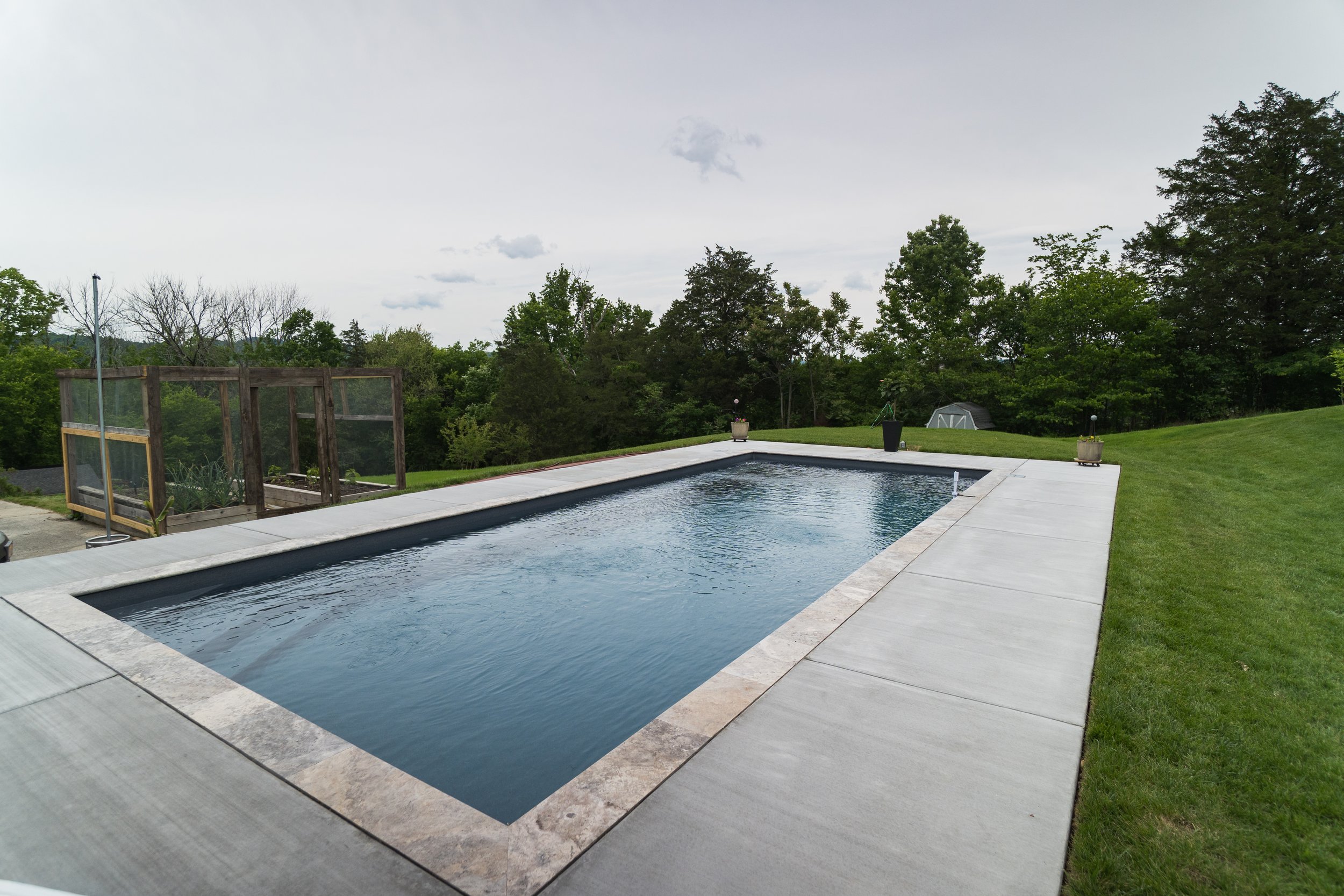 An outdoor backyard scene with a swimming pool surrounded by concrete and green grass, trees and cloudy sky in the background, and a small garden area with a wooden frame on the left side.