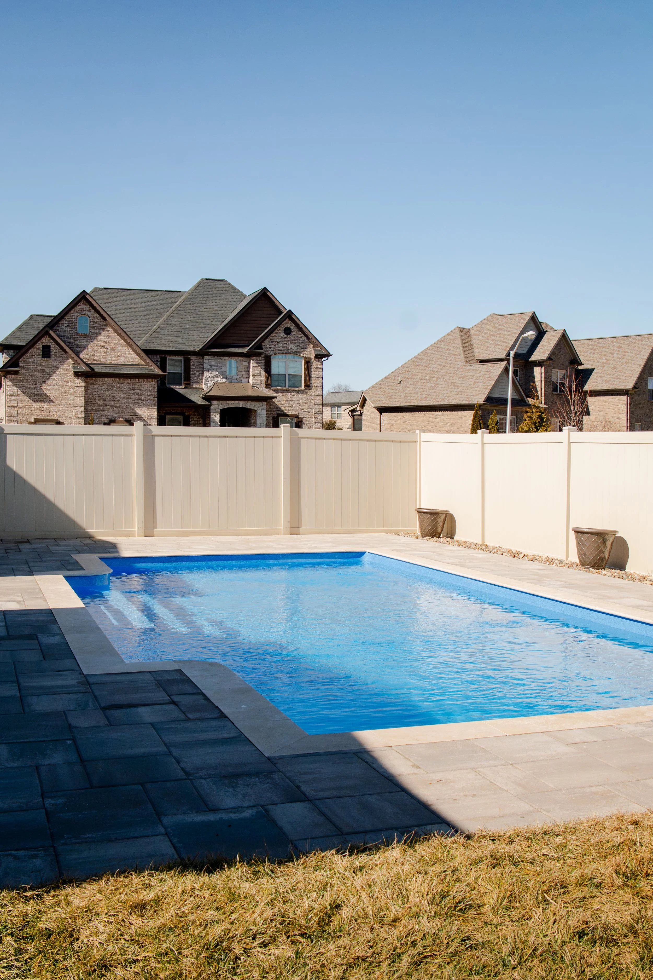 A backyard with a swimming pool, a white fence, and neighboring brick houses under a clear blue sky.