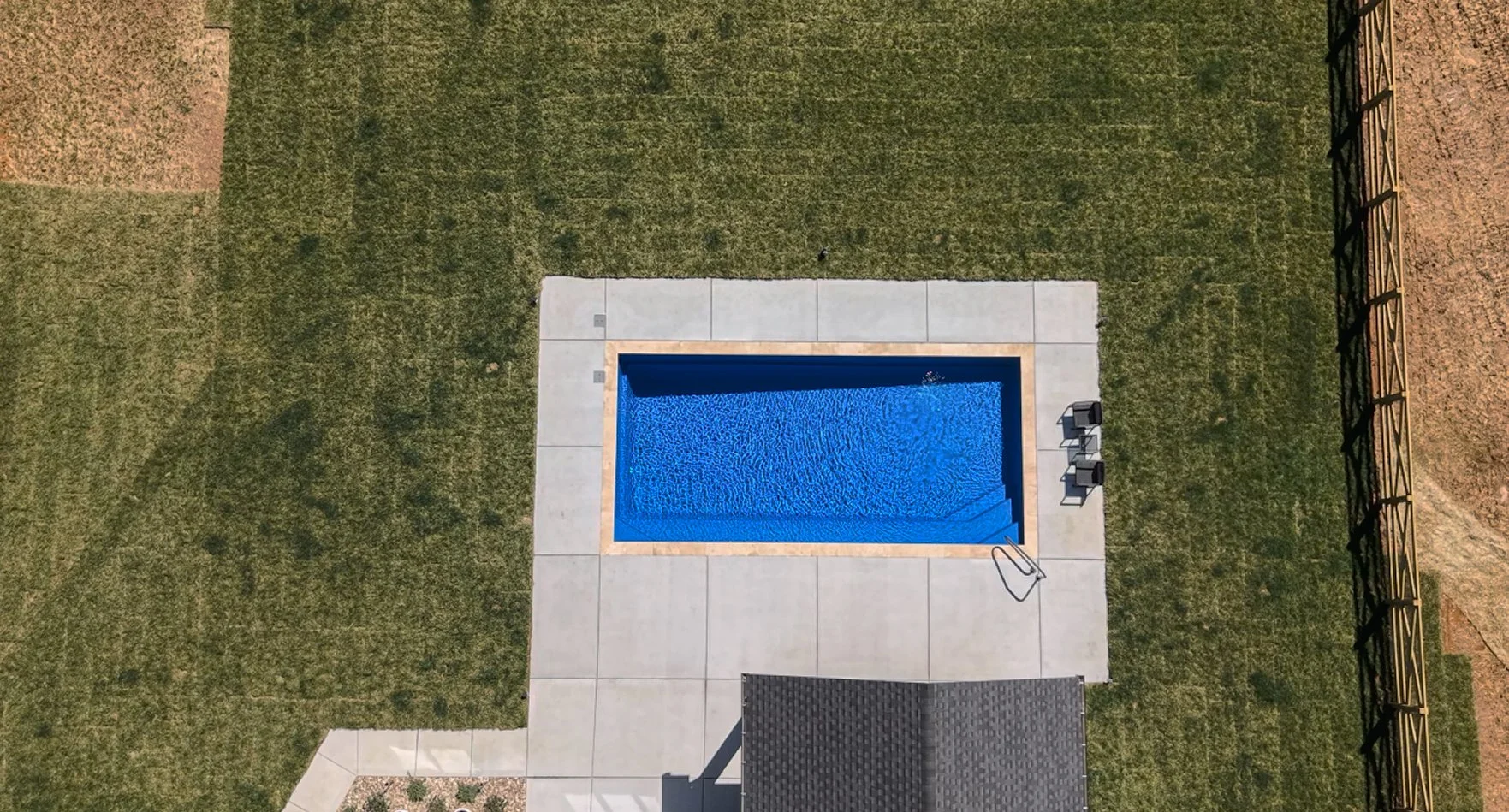 An aerial view of a rectangular swimming pool with blue water, surrounded by a concrete deck and green grass area. There are two lounge chairs and pool steps at one end.