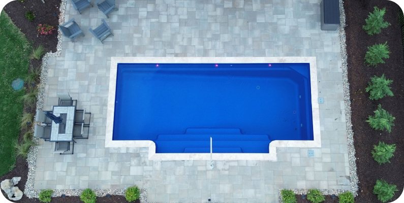 A rectangular outdoor swimming pool with steps entering from one side, surrounded by a paved patio with outdoor furniture and landscaping, viewed from above.