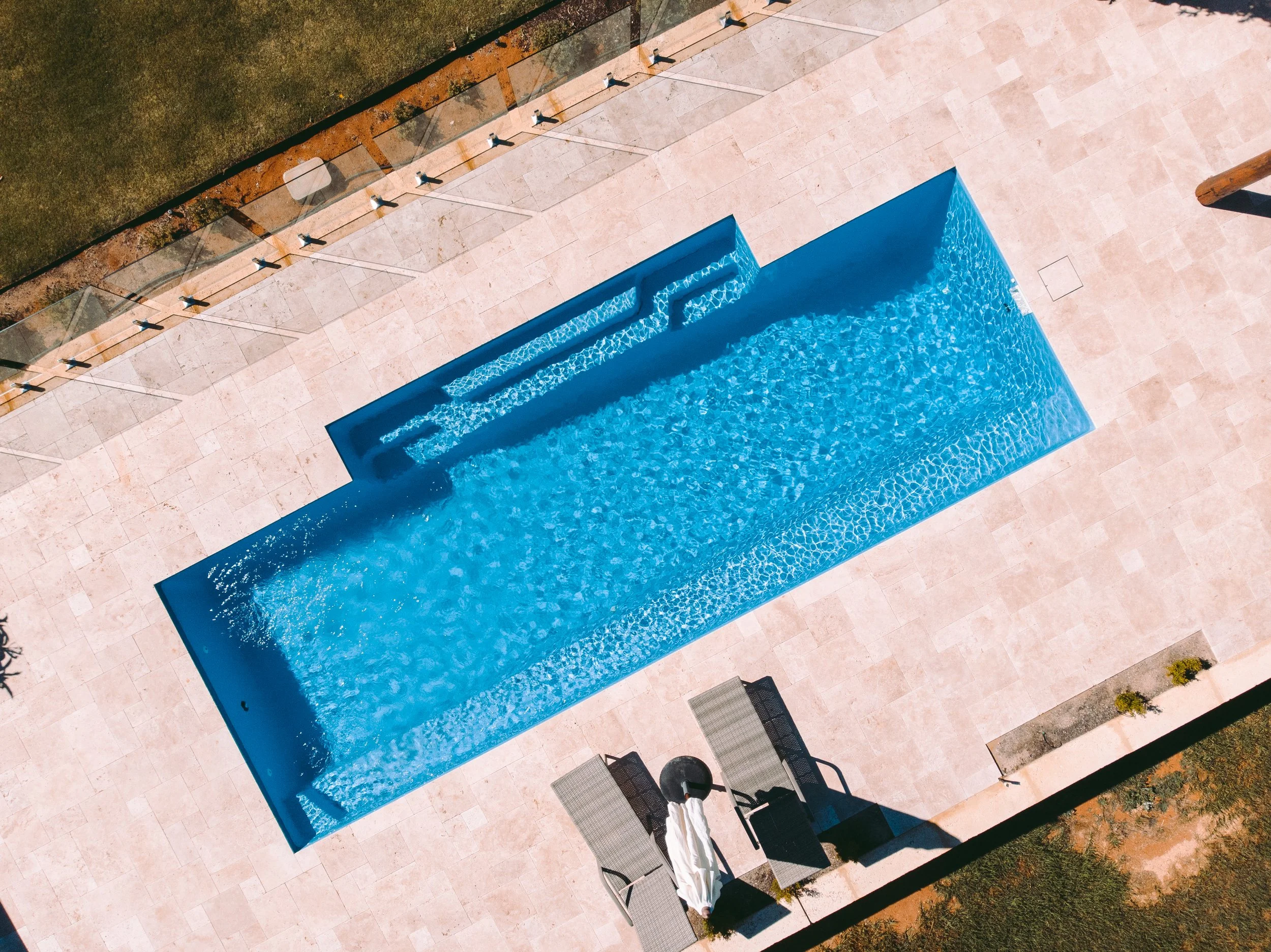 Aerial view of a swimming pool with steps, surrounded by a tiled patio, lounge chairs, and landscaping.