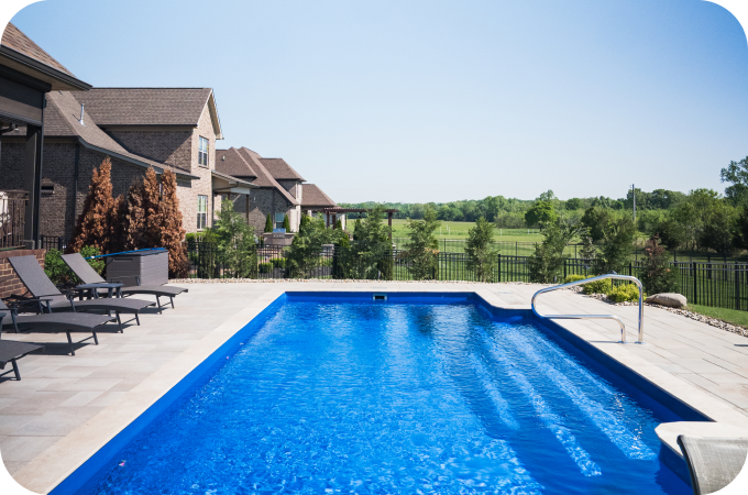 Residential backyard with a swimming pool, lounge chairs, and a view of a grassy field and trees in the distance.