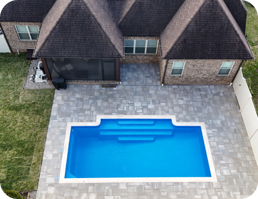 A backyard with a rectangular swimming pool surrounded by gray paving stones, next to a brick house with a sloped roof.