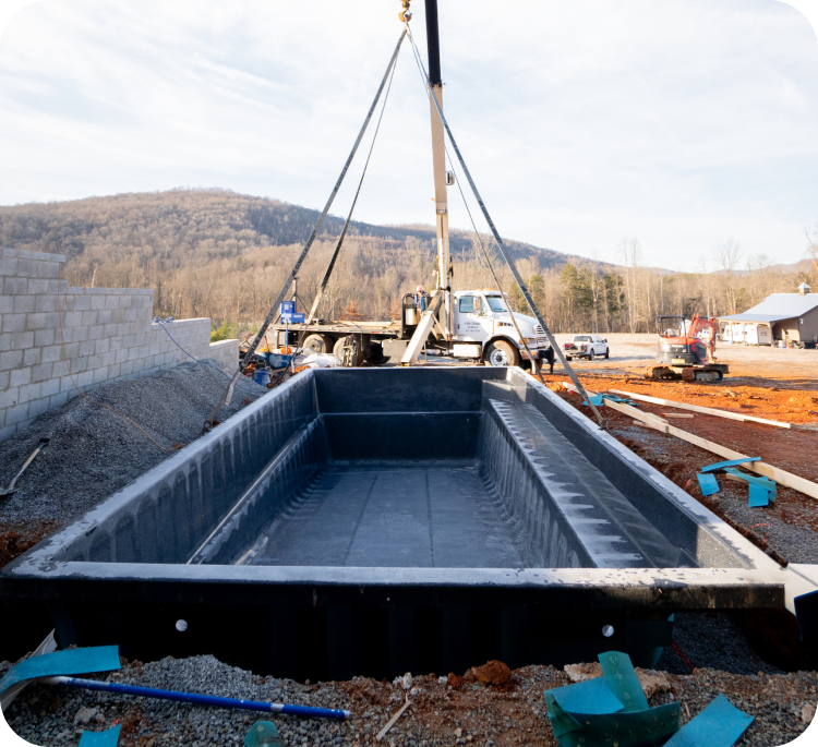 Construction site with a large black concrete mold or basin being lifted by a crane, with construction vehicles and equipment in the background, and a partially built wall to the left.
