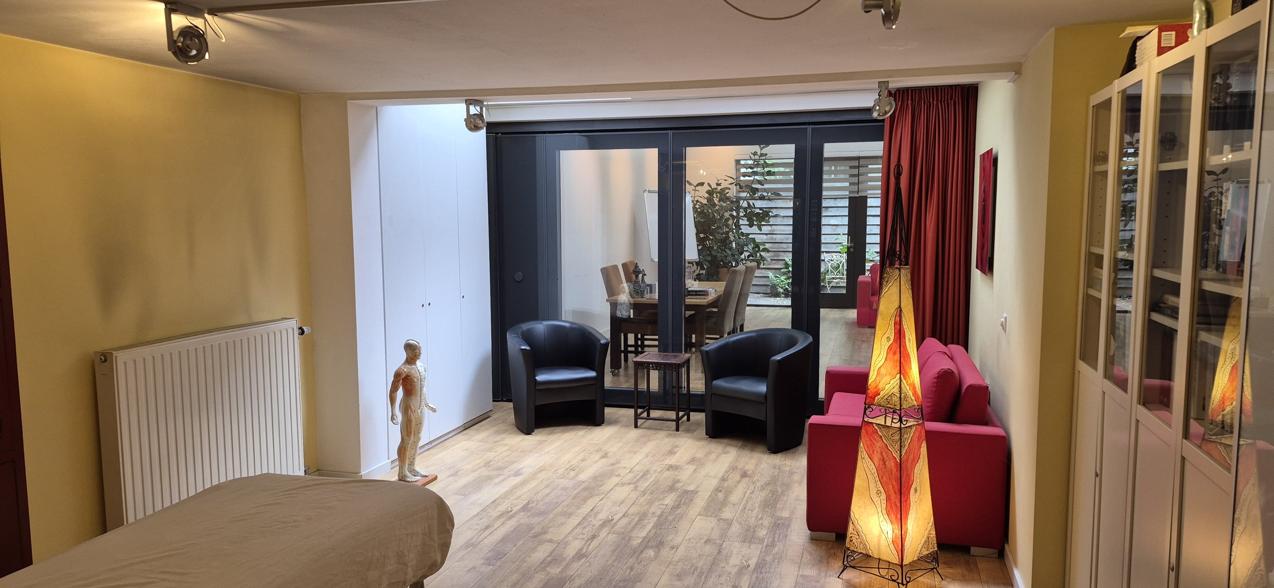 Gezellige woonkamer met zwarte fauteuils, rode bank, kleurrijke vloerlamp, glazen deuren naar de eetkamer en een vitrinekast.