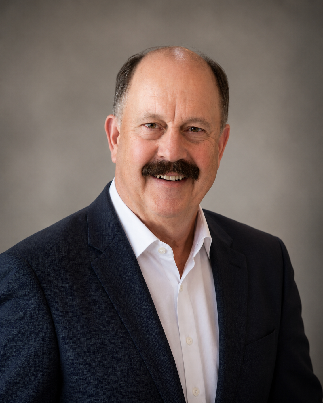 A smiling man with a mustache in a dark suit and white shirt against a gray background.