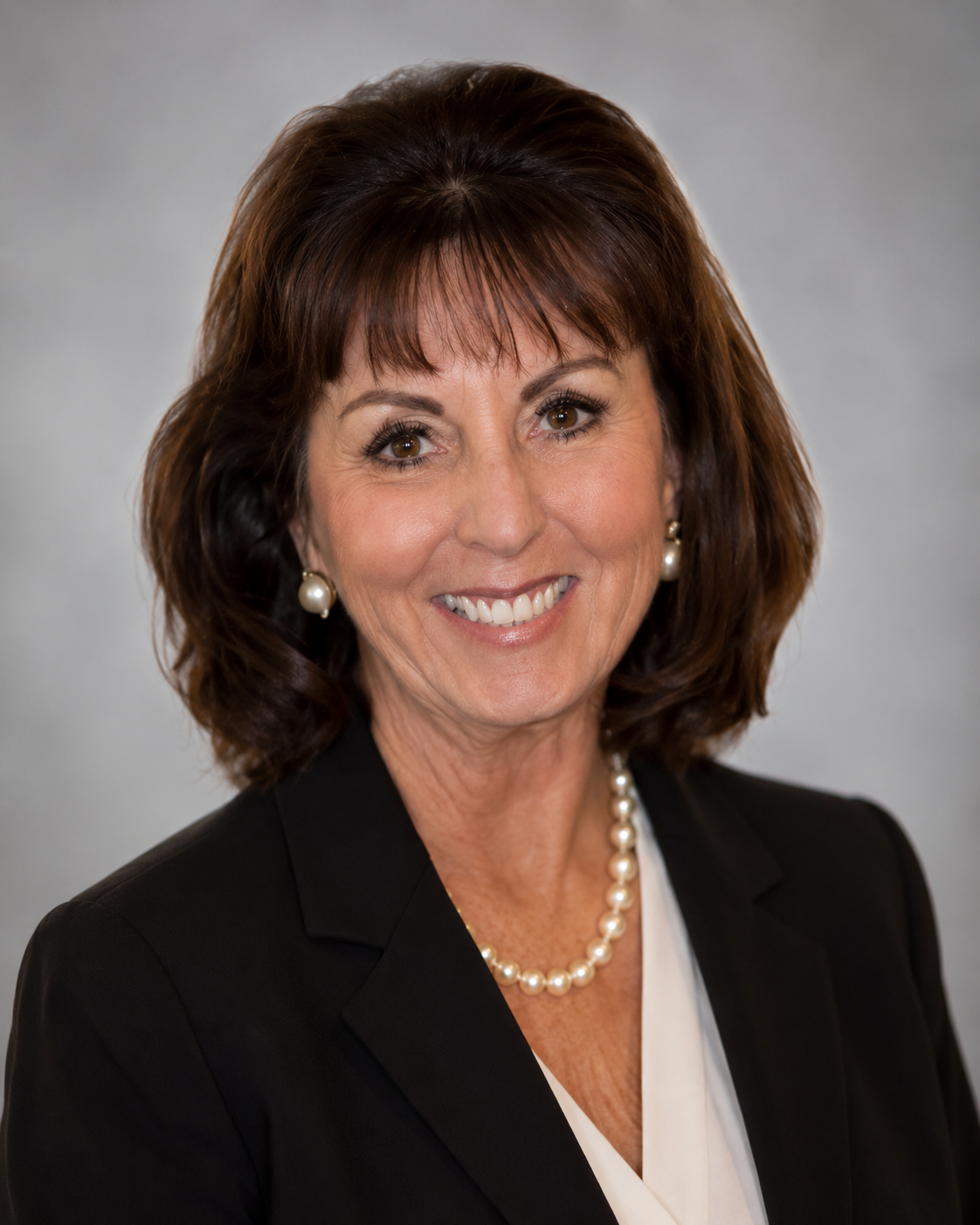 A professional woman with brown hair and brown eyes, wearing a black blazer, white blouse, pearl necklace, and pearl earrings, smiling against a gray background.